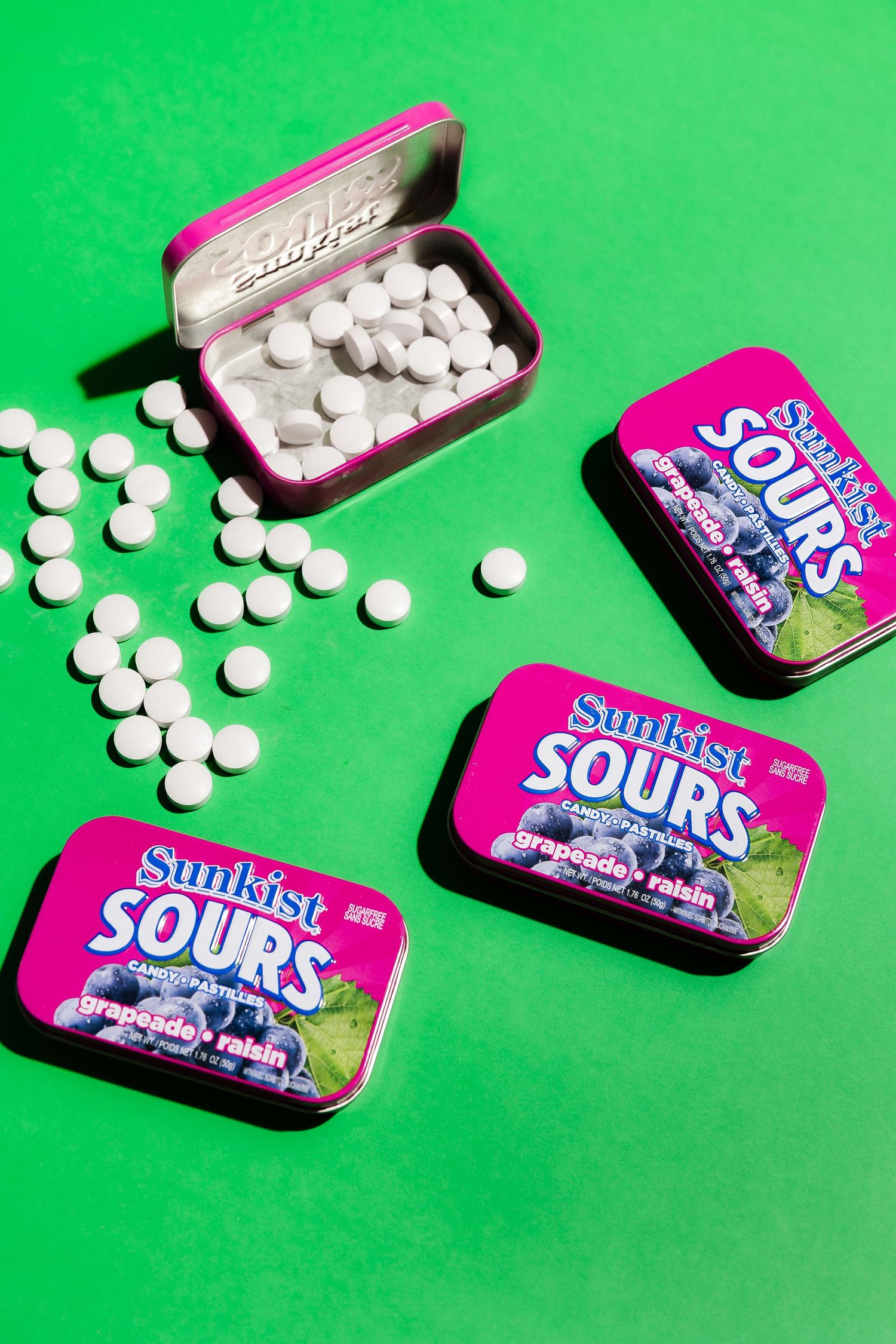 White sour candies scattered around pink Sunkist tins on vibrant green surface.