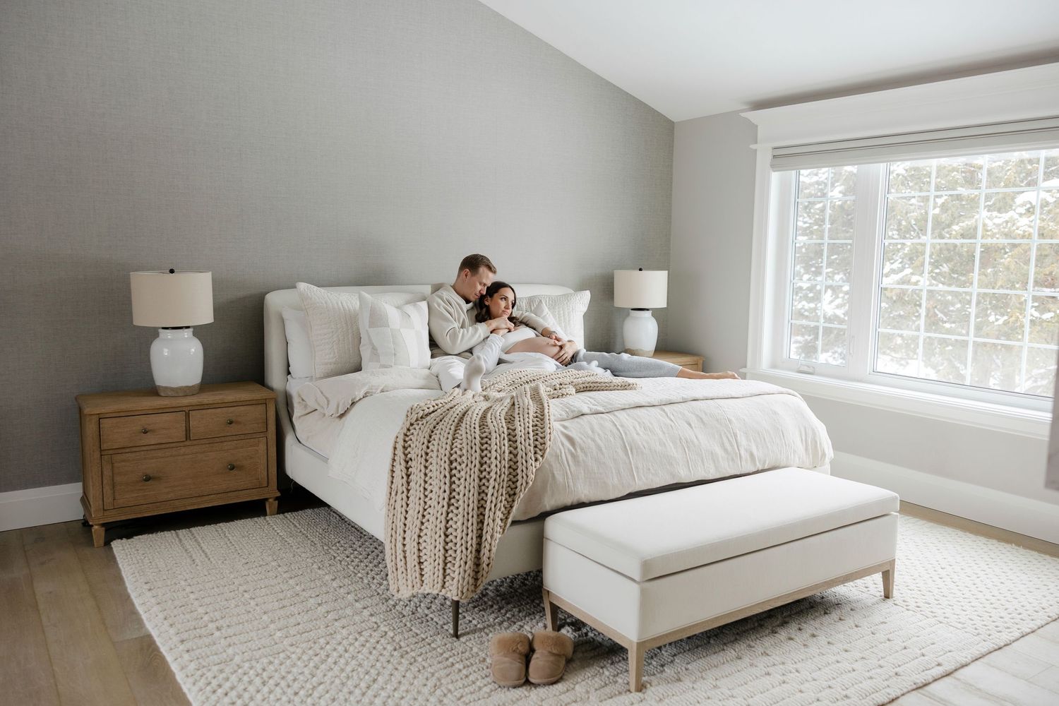 Cozy bedroom scene with neutral tones, featuring plush bedding and natural light from large windows.