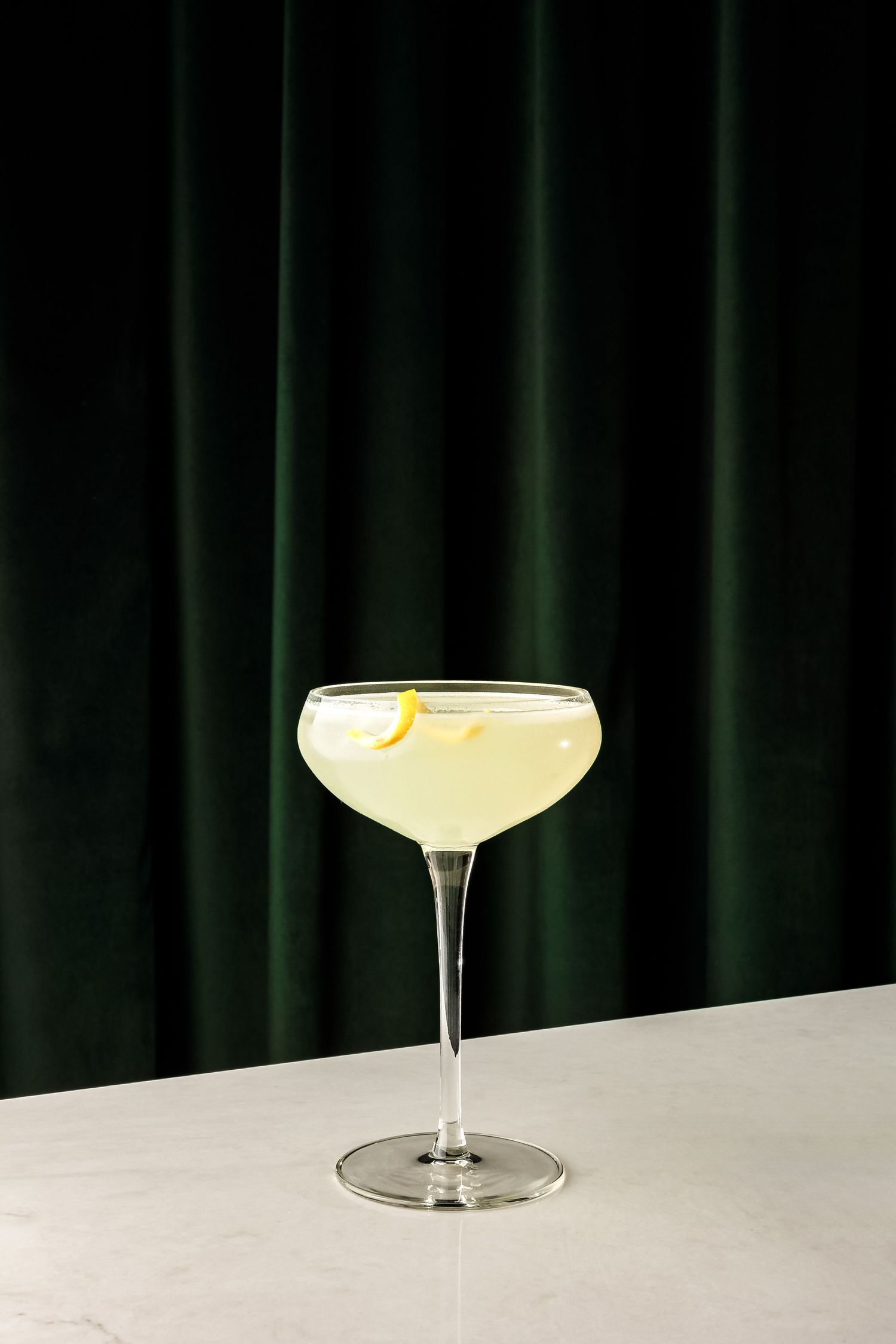 Classic coupe glass filled with light colored cocktail against dark green backdrop.