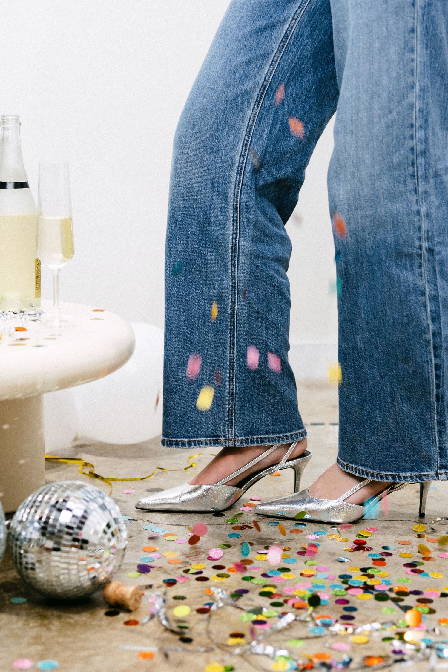 Jeans and silver heels surrounded by colorful confetti and disco ball at party.