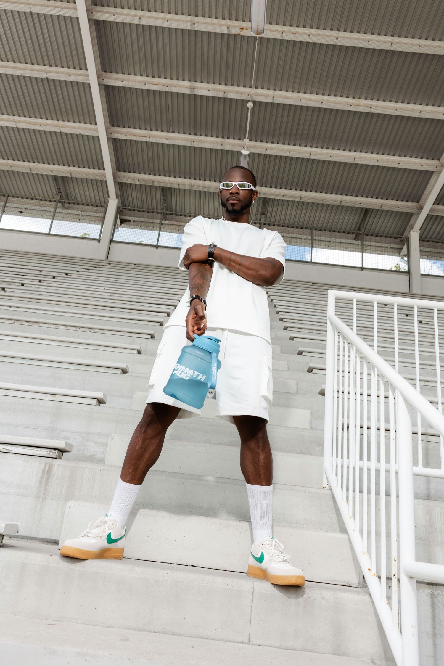 Athletic figure in white outfit and sunglasses posing dynamically on concrete stadium steps with blue water bottle.