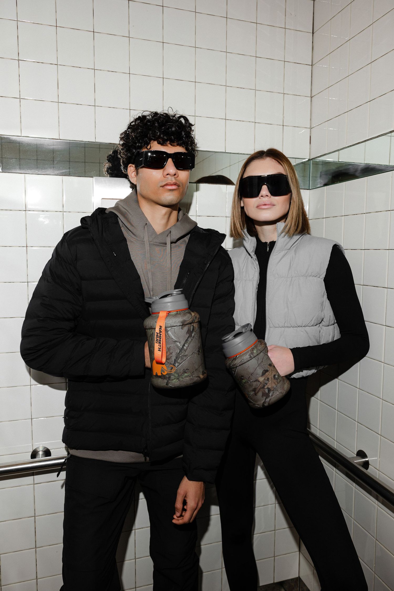 Two people in black outfits and sunglasses pose against a white tiled wall while holding drinks.