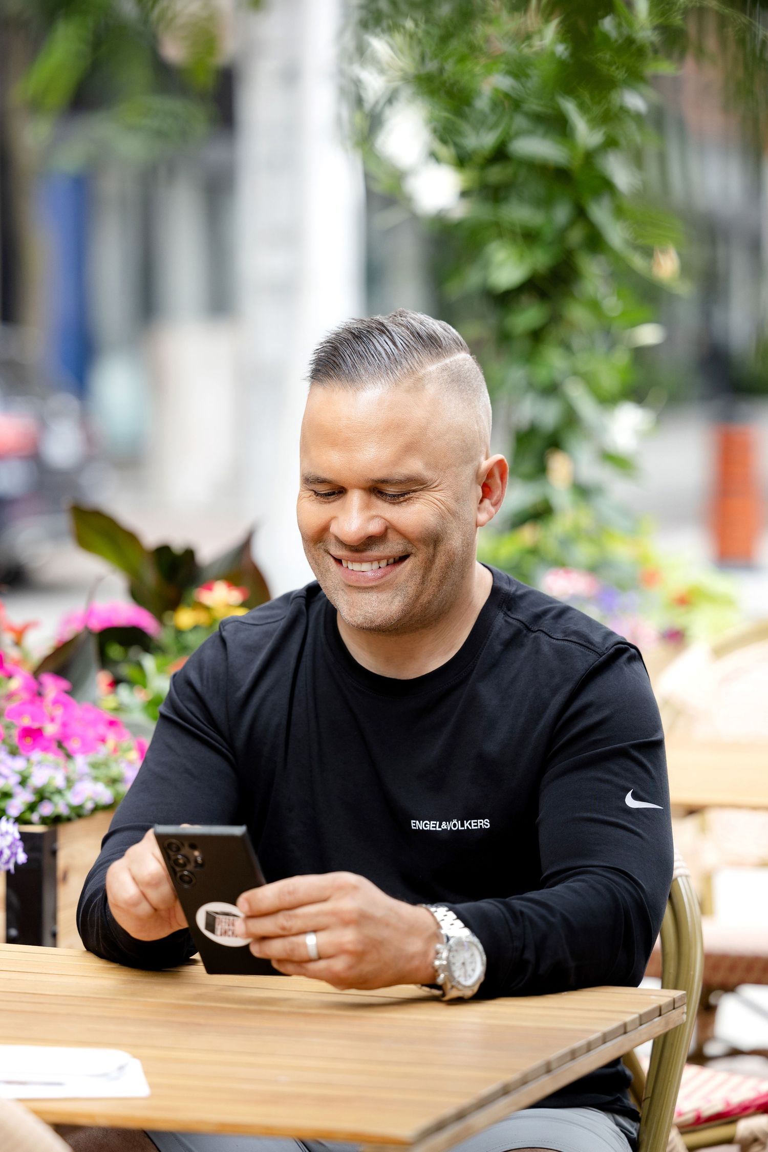 Person in black shirt smiling while looking at smartphone at outdoor cafe table.
