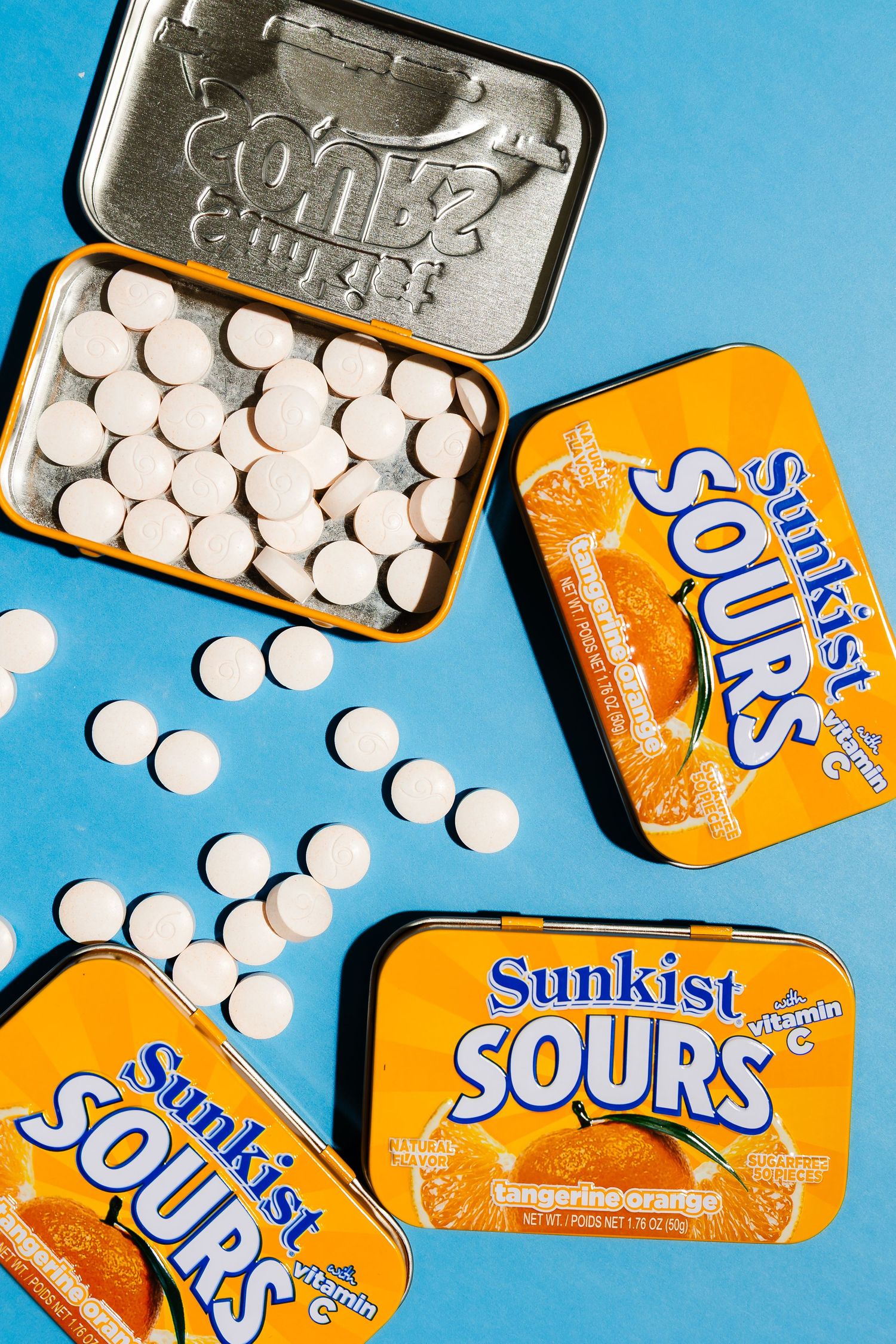 Orange Sunkist Sours candy tins scattered with white tablets on blue background.