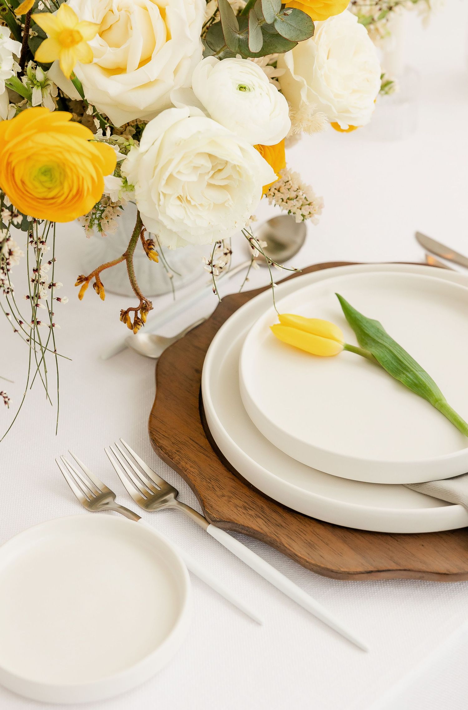 Elegant place setting with white plates on wooden charger surrounded by yellow and white roses.