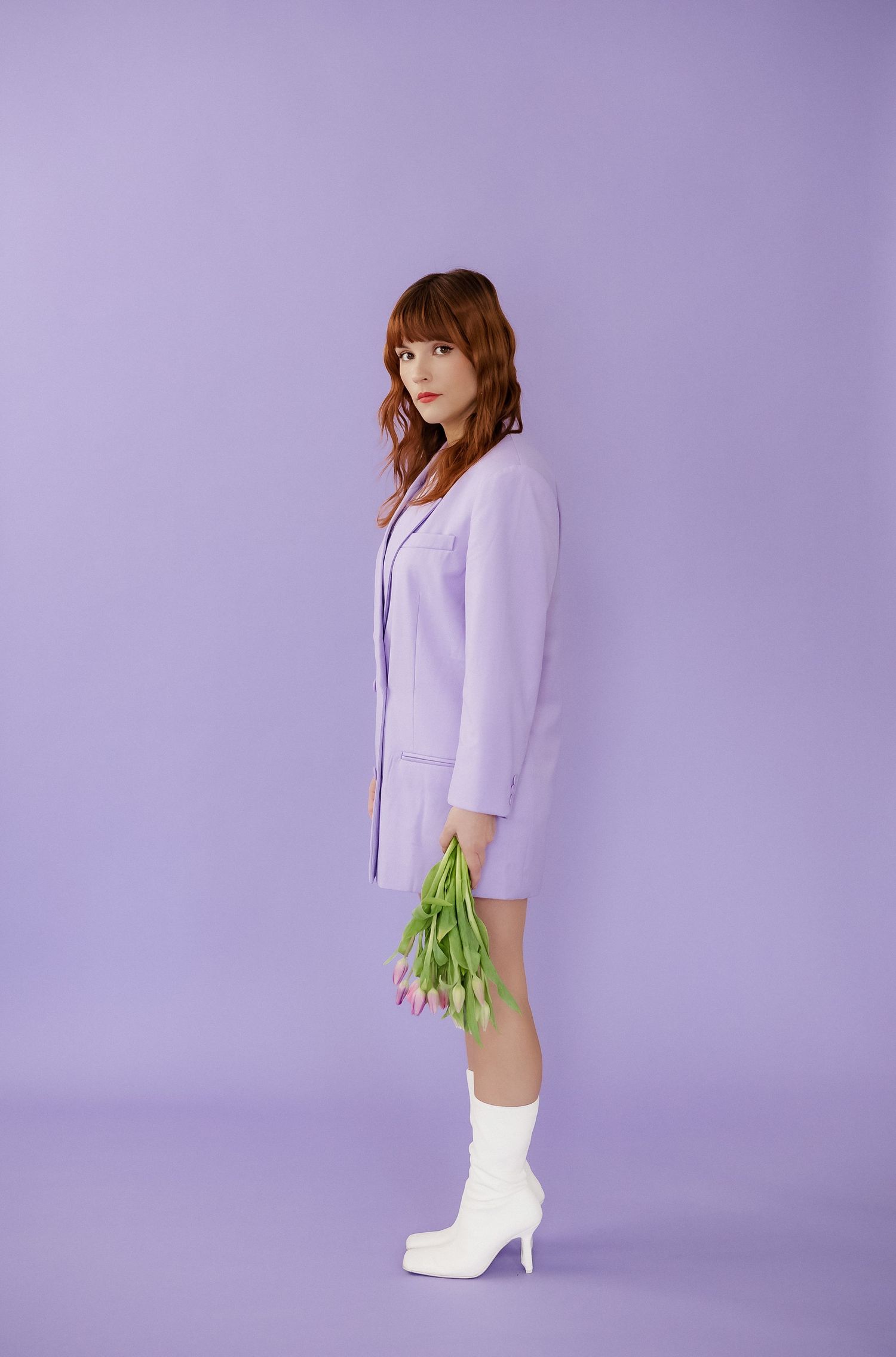 Stylish figure in lilac blazer and white boots stands against purple background.