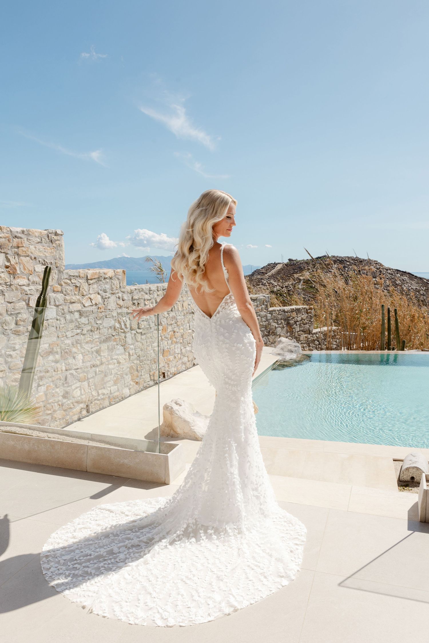 A bride in a white lace gown stands on a terrace overlooking a stunning coastal view.