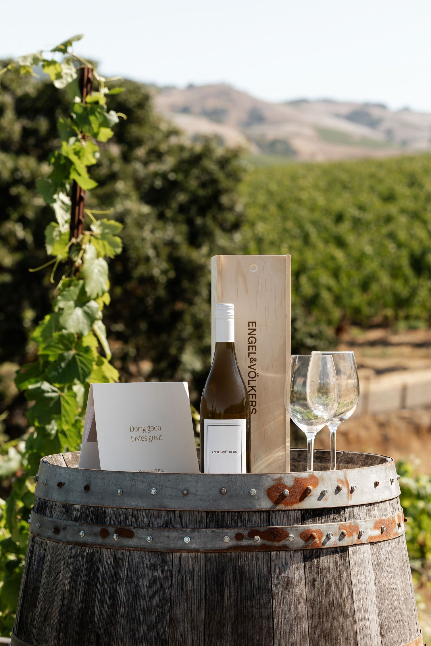 Wine bottles and branded merchandise displayed on rustic wooden barrel in vineyard.