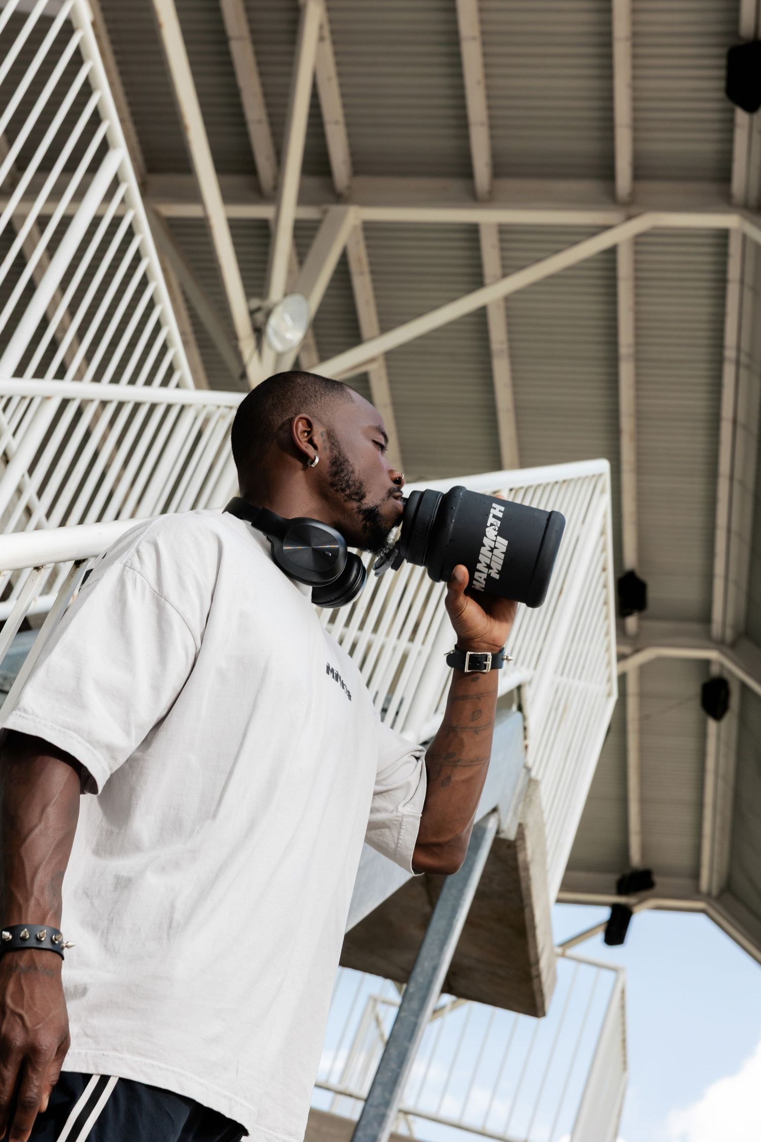 Athletic person in white shirt drinking from black water bottle near stairs.