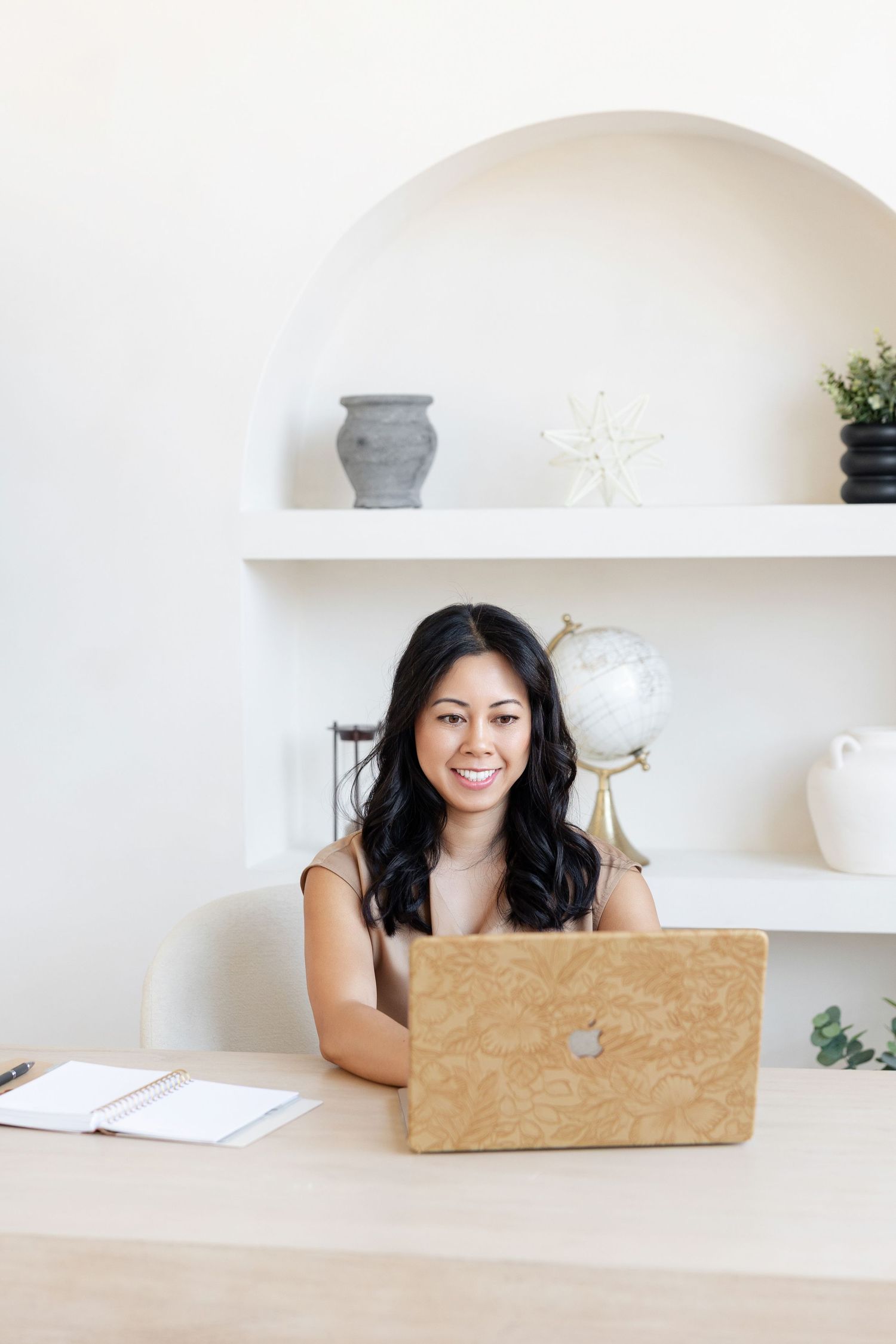 Person using laptop at bright white desk with decorative shelving and accessories.