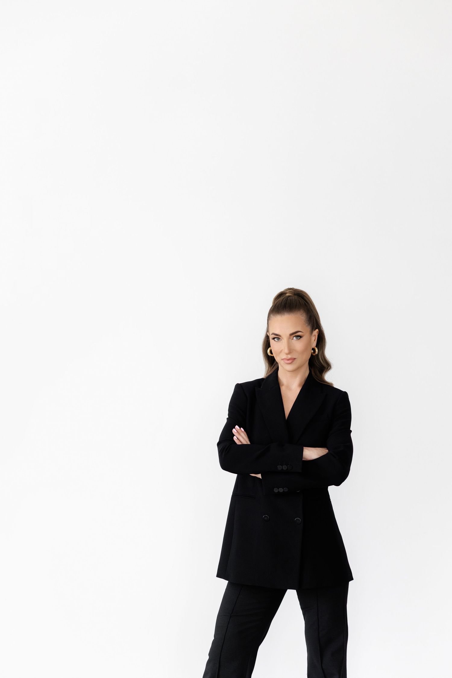Woman in black power suit poses with crossed arms against minimalist white backdrop.