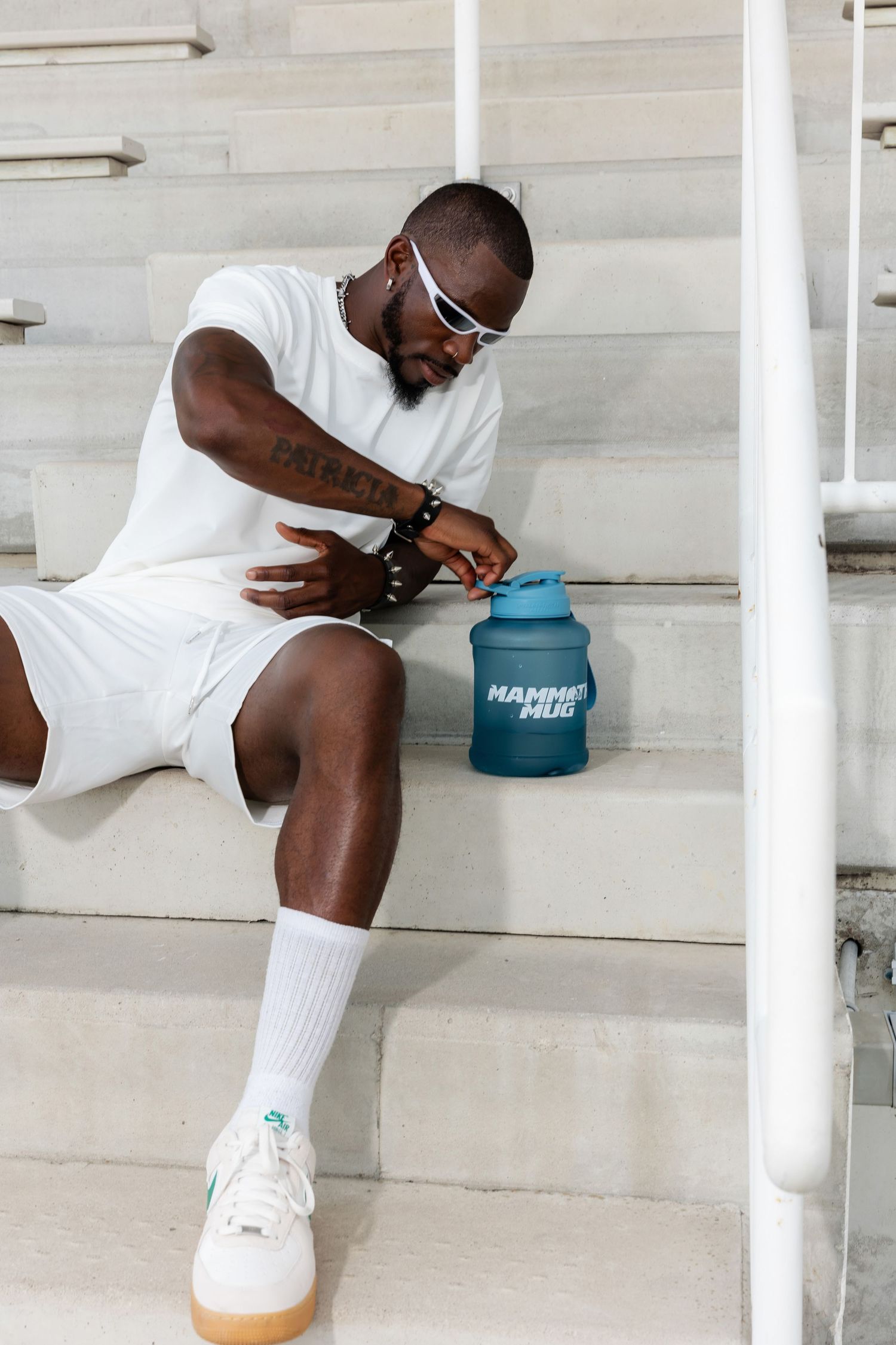 Person in white outfit and sunglasses sitting on bleachers with blue water bottle and white sneakers.