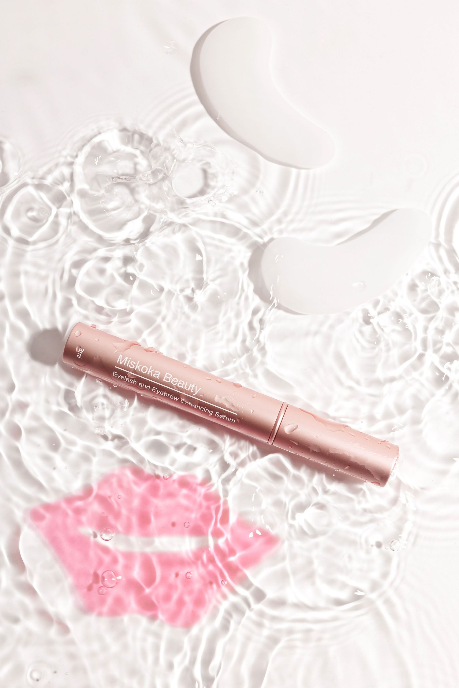 Rose gold beauty pen on white surface with pink lip prints and water ripples.