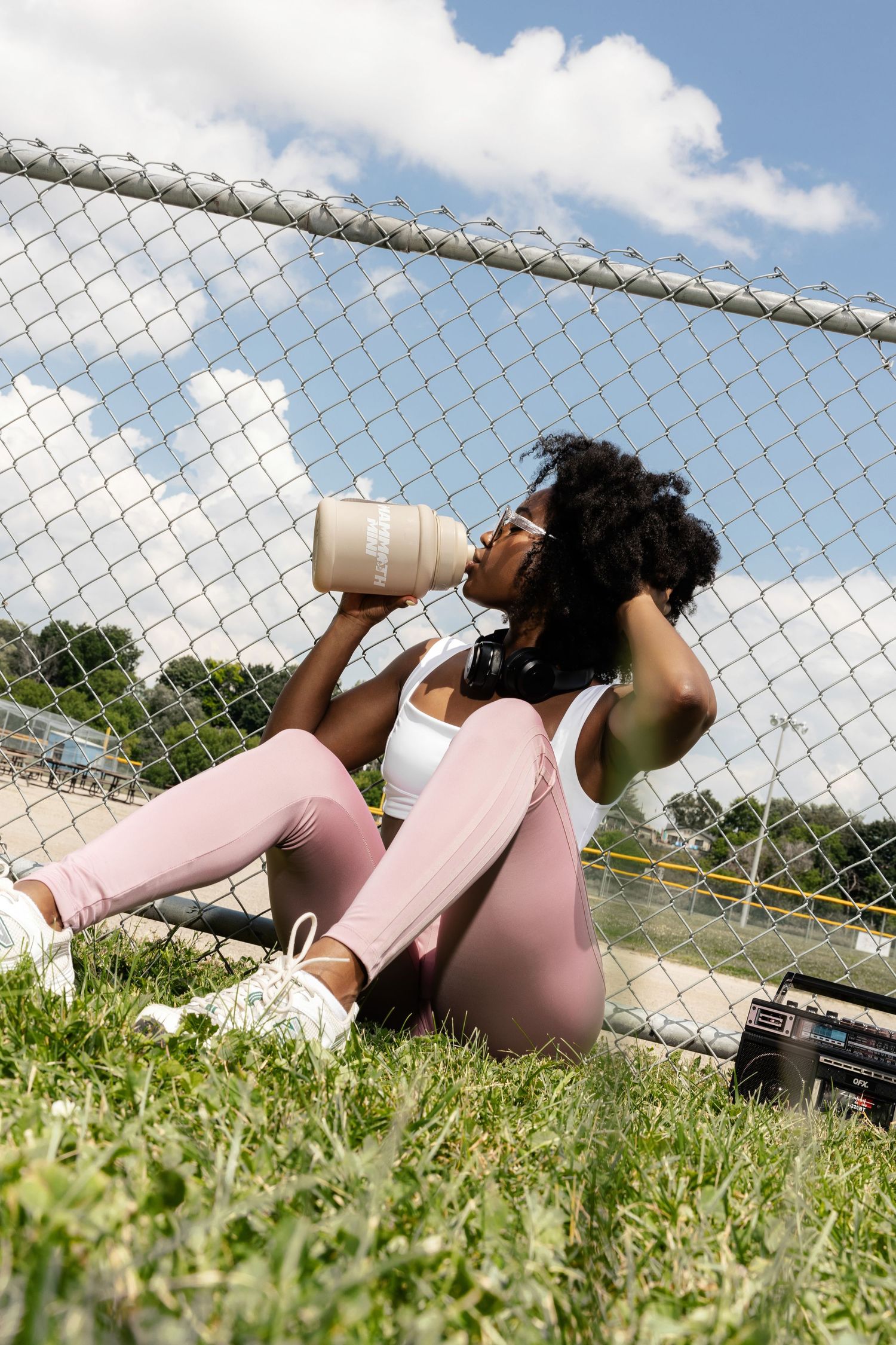 Person in pink athletic wear lying on grass near chain link fence with water bottle.