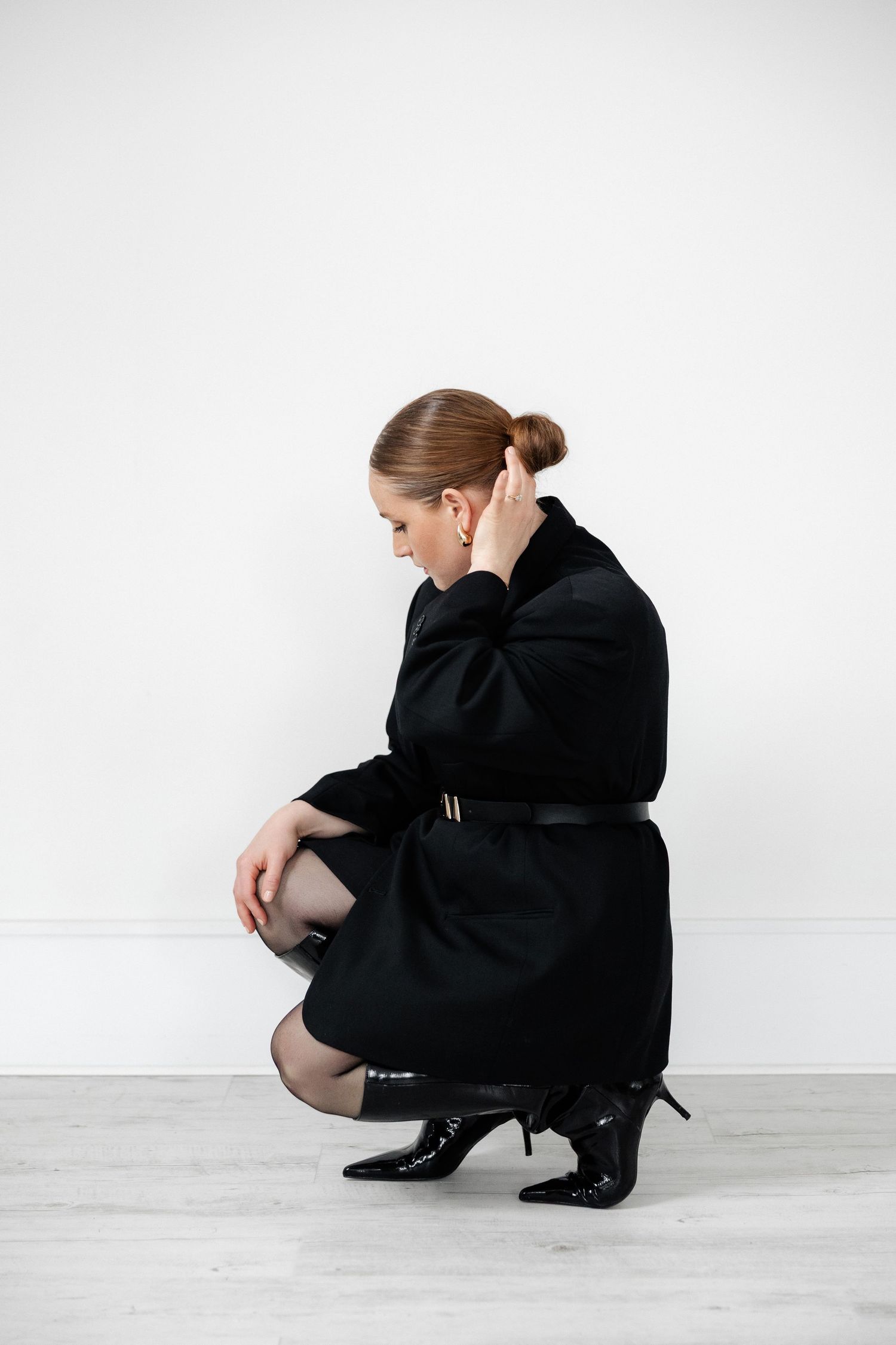 Figure in oversized black coat crouches while adjusting hair against white backdrop.