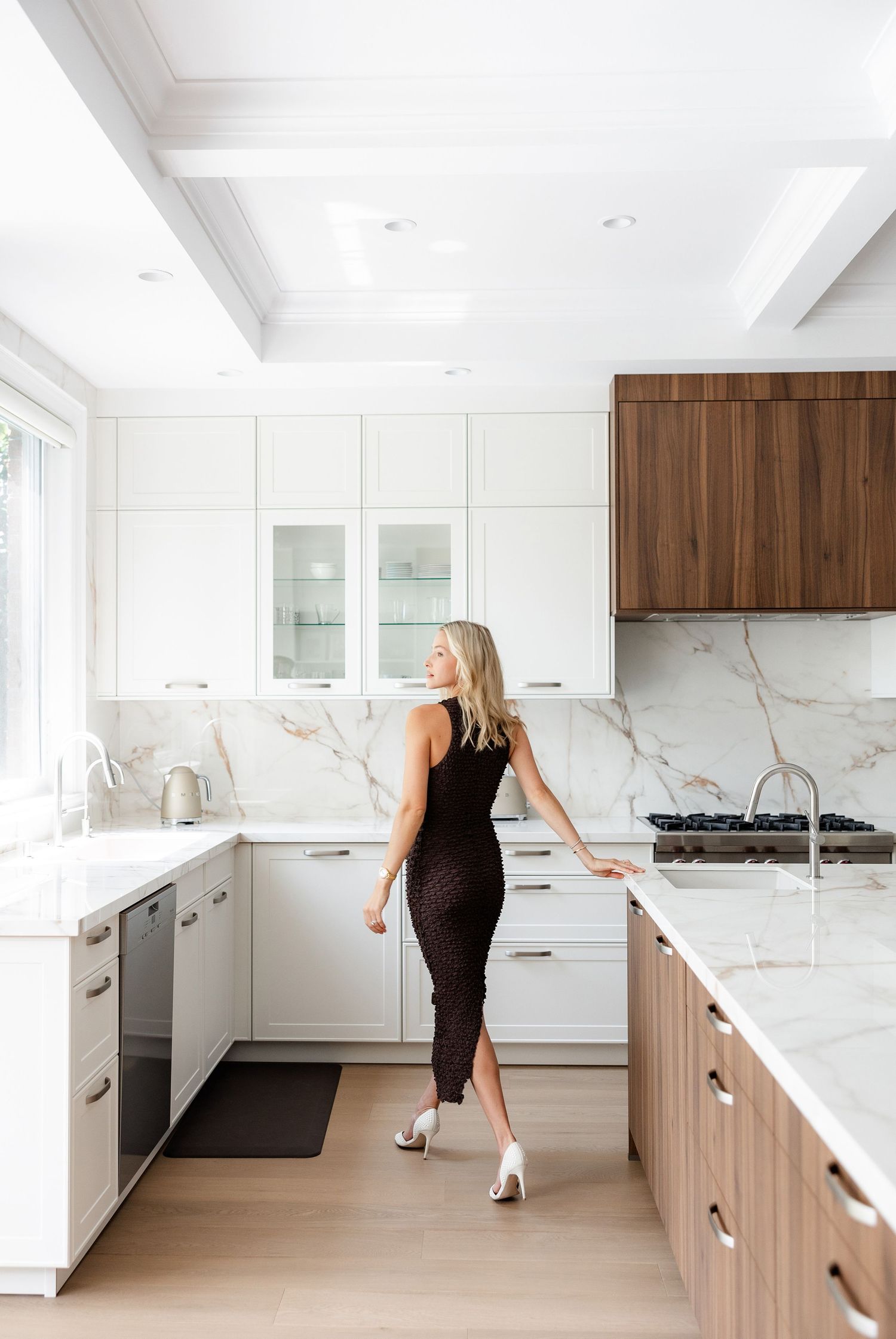 Woman in black dress explores modern white kitchen with marble countertops.