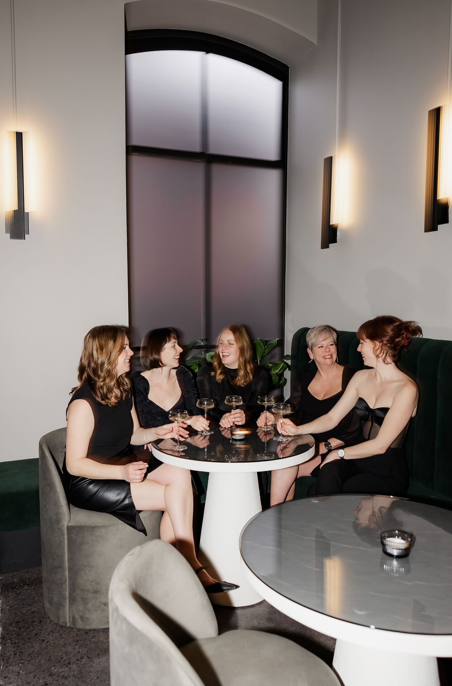A group in black dresses enjoying drinks and conversation at a modern lounge with wall sconces and curved seating.