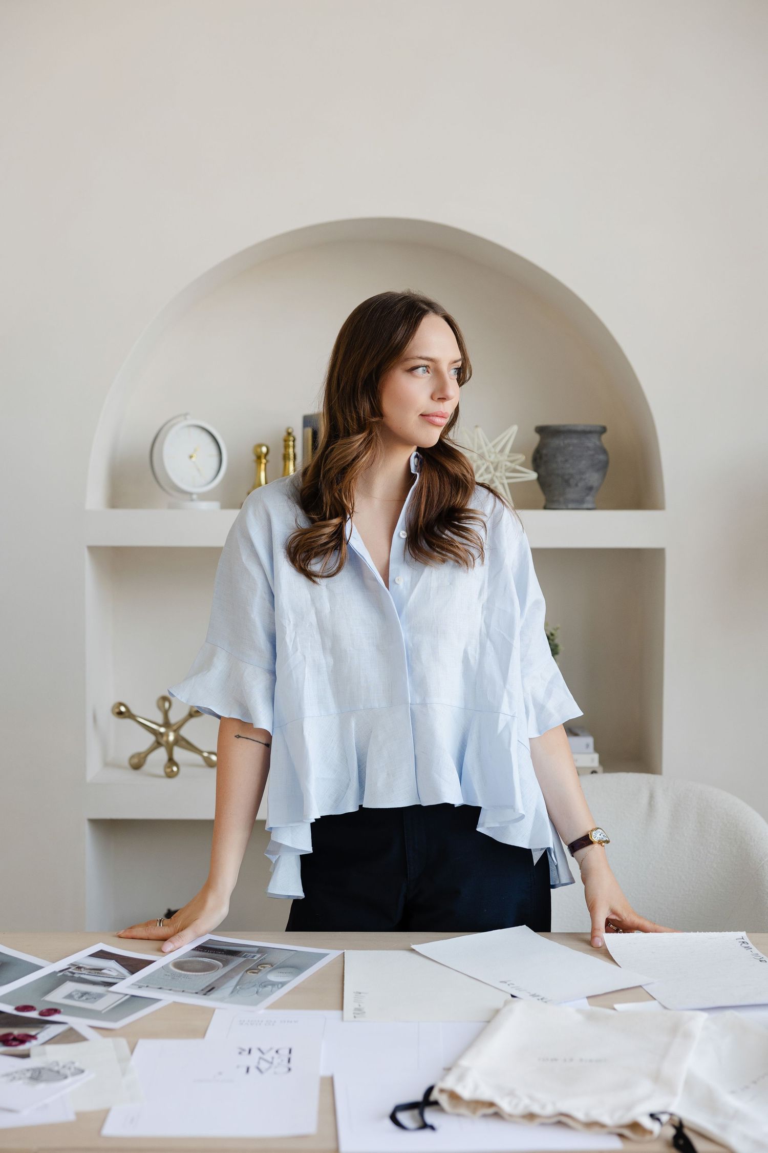 Designer in light blue blouse reviews materials at workspace with built-in shelving.