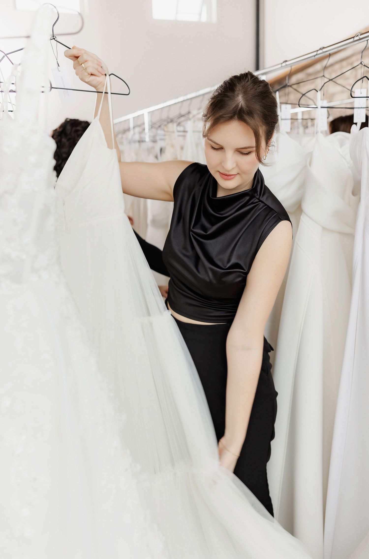 Person in black examining white wedding dresses on hangers in a bridal boutique.