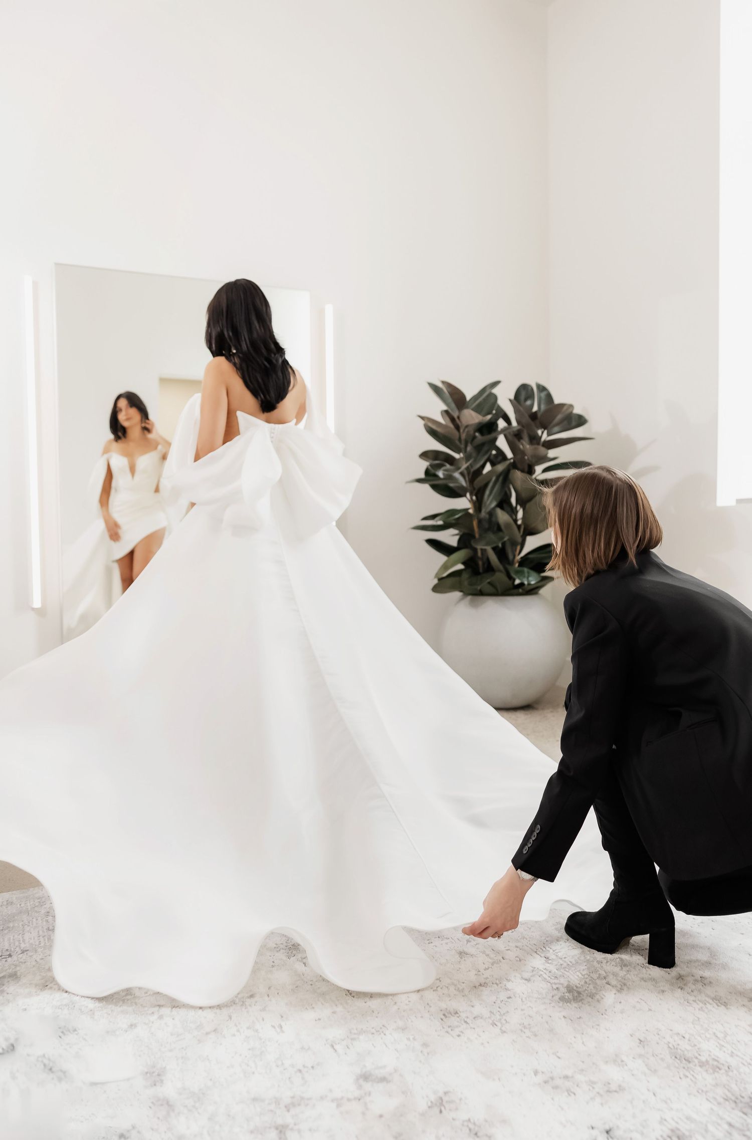 A bridal gown fitting session showing a stylist adjusting the train of an elegant white wedding dress in a bright studio.