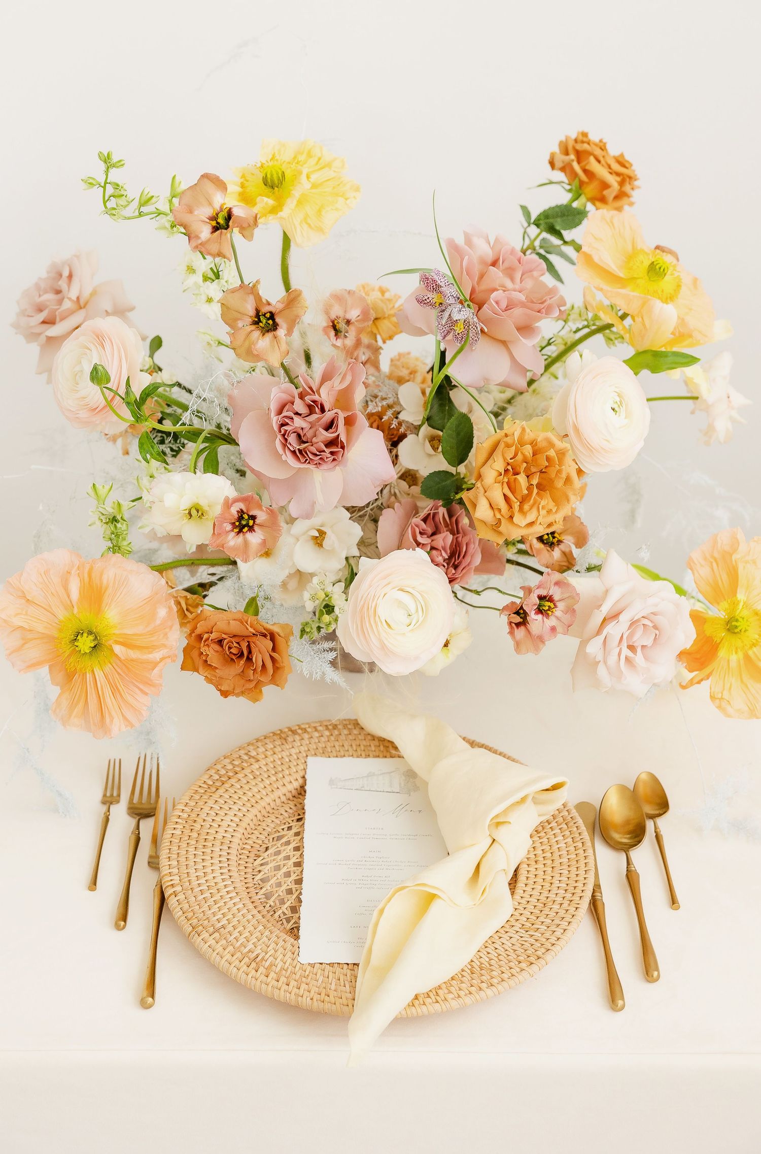 Table setting with woven charger plates, gold cutlery and romantic floral centerpiece in peach and yellow.