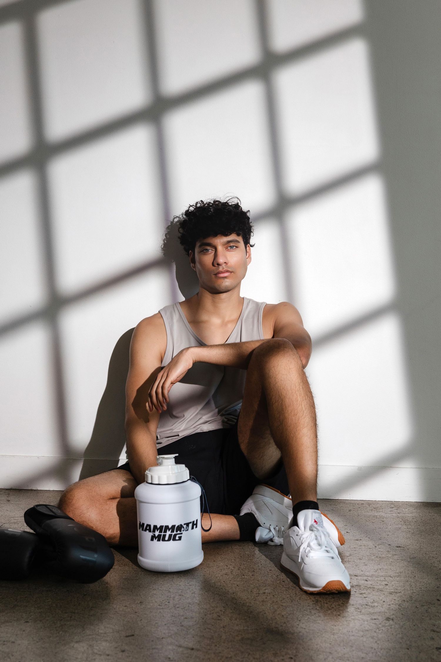 Athletic model in tank top sits casually with white branded container and sneakers.