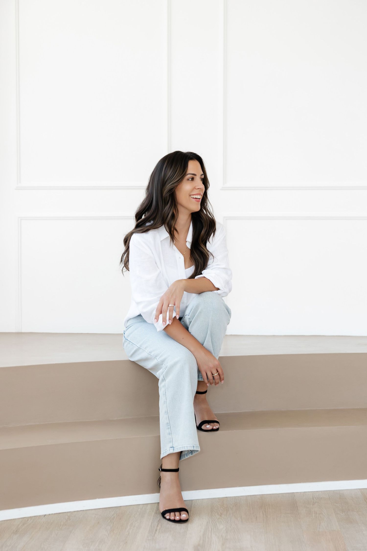 Woman in business casual attire poses on beige steps with white wall background.