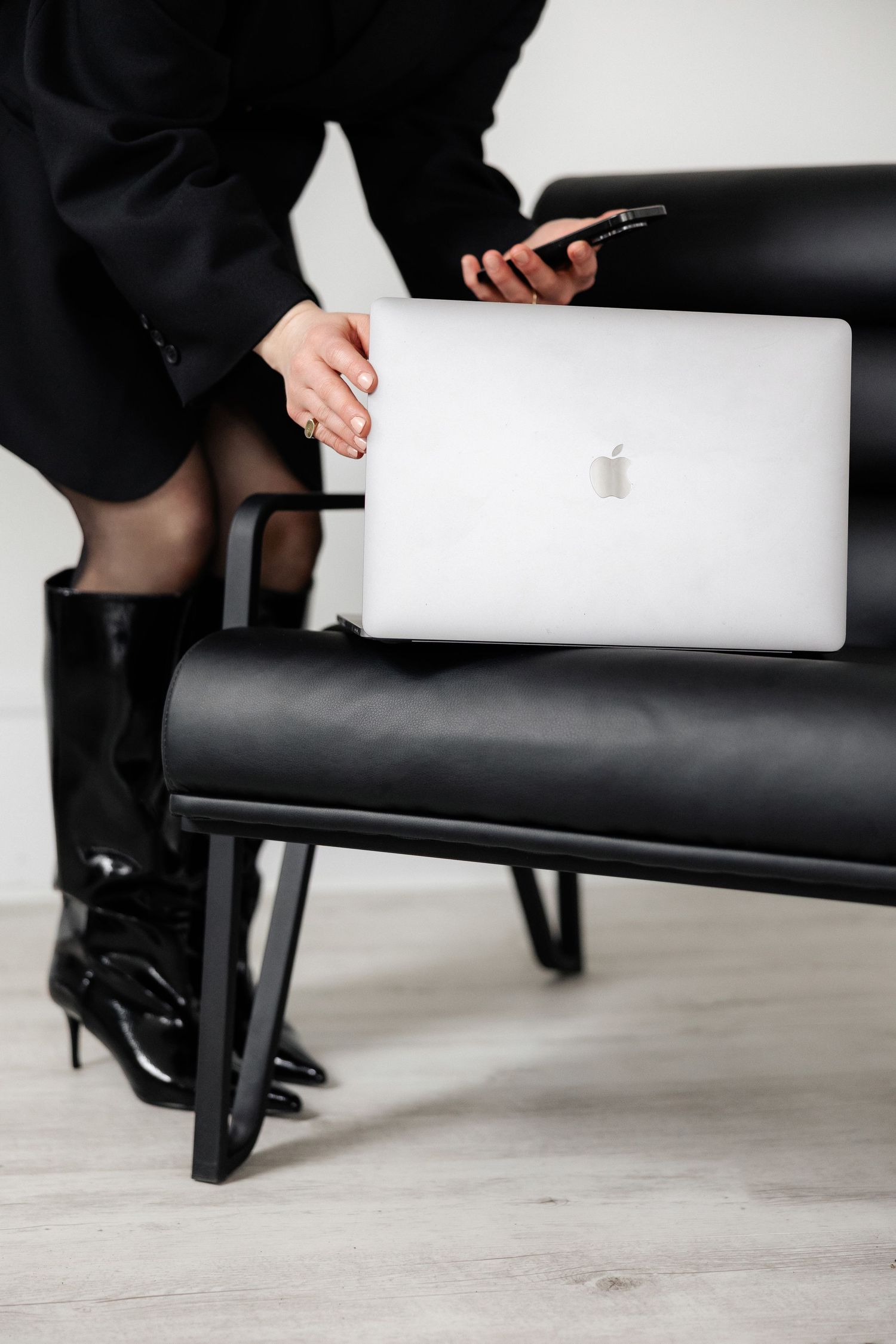 Person in black outfit with boots using MacBook and phone on leather chair.