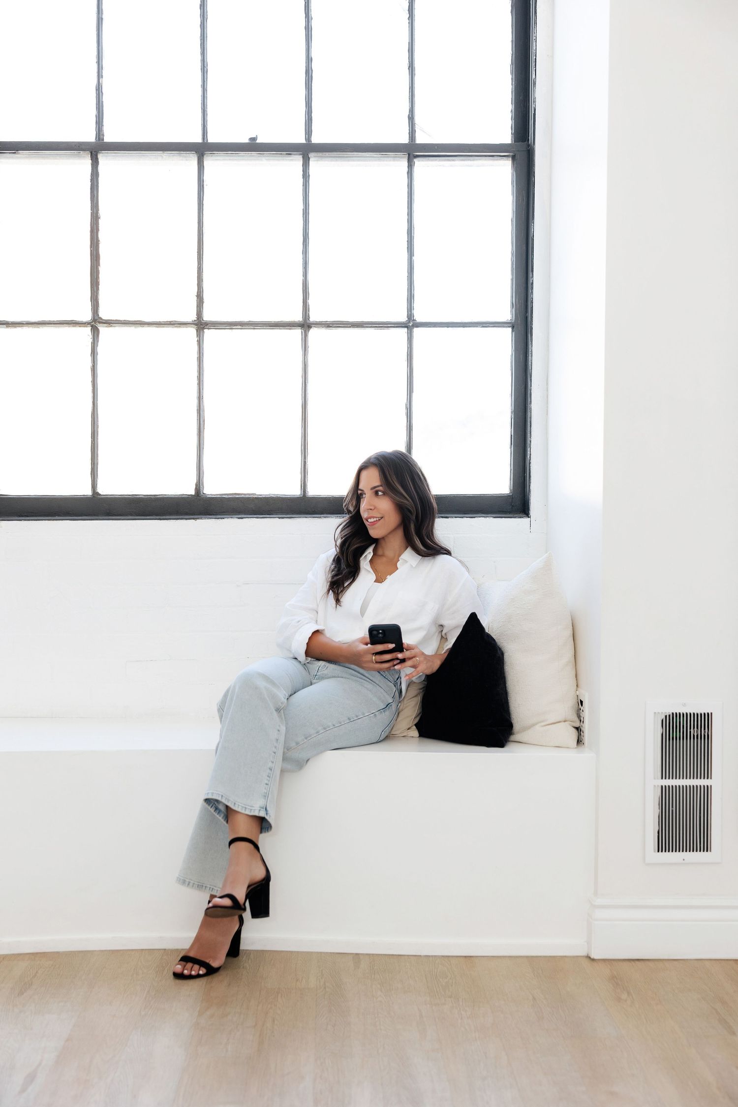 Person relaxing on white couch while using phone in bright industrial space with windows.
