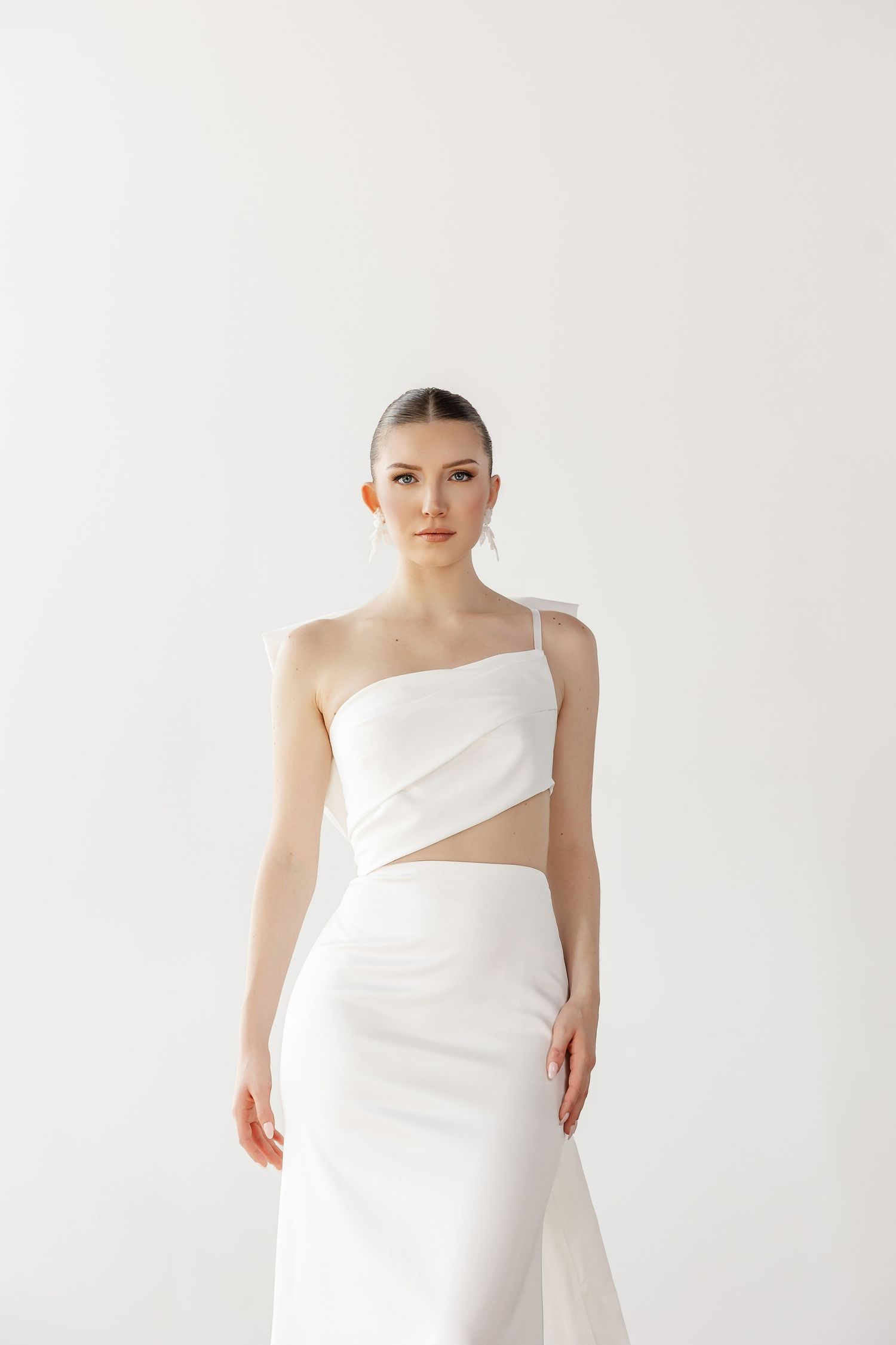 Modern minimalist white bridal gown with asymmetrical neckline and cutout detail shown in studio.