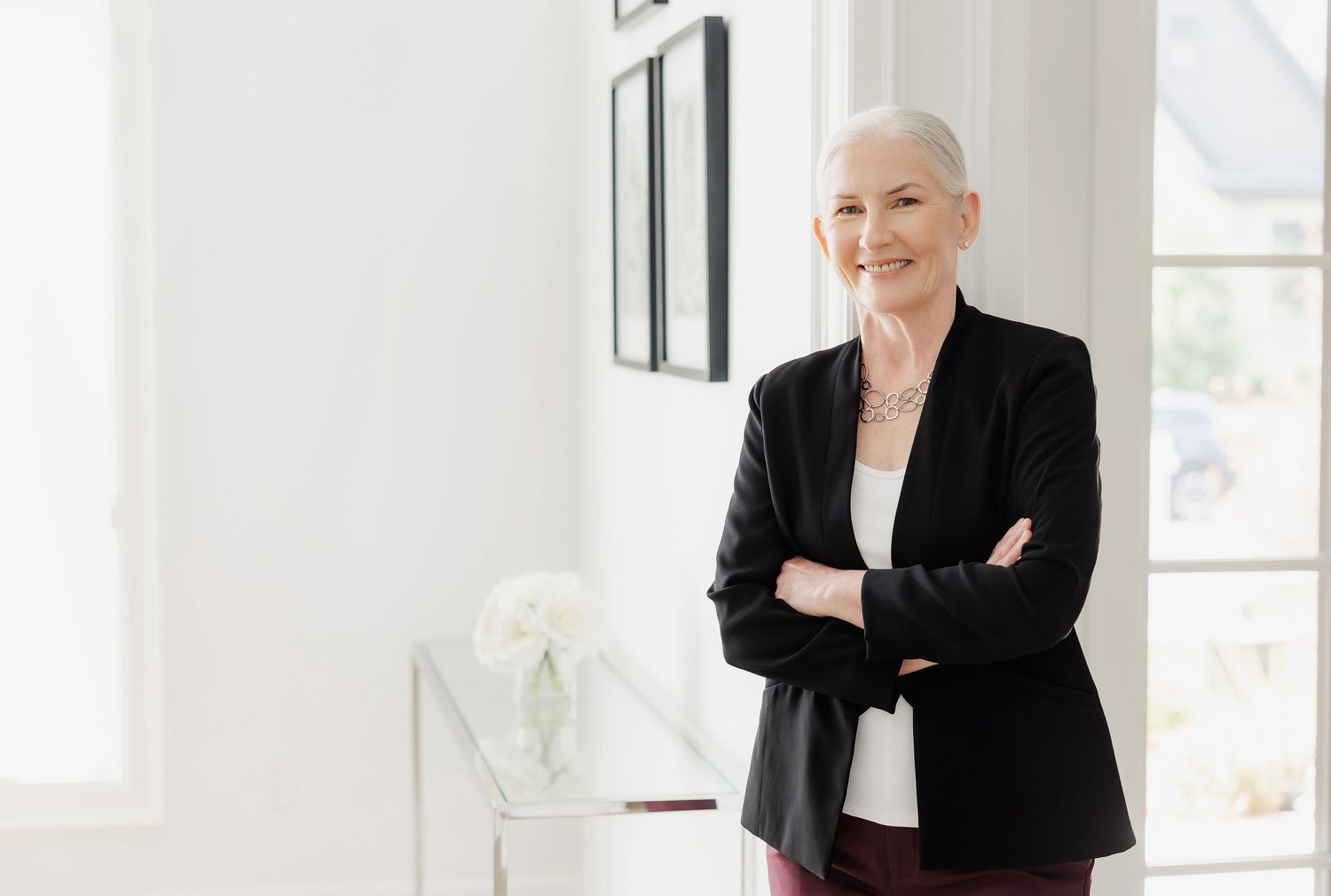 Executive in black blazer stands confidently with arms crossed in bright modern office space.