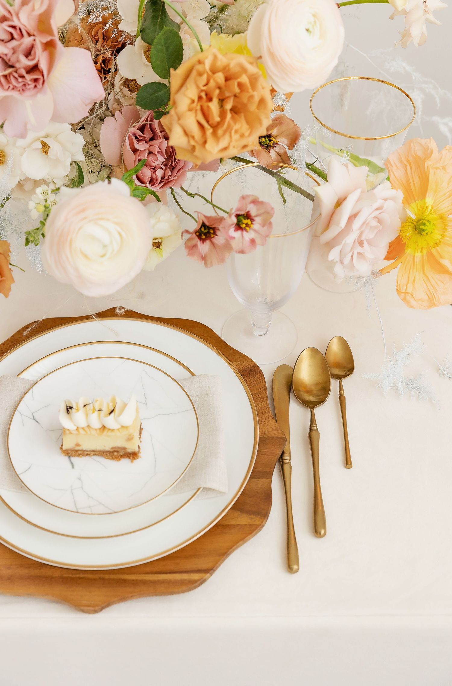 Elegant place setting with wooden charger plate, white dinnerware and gold accents under floral arrangement.