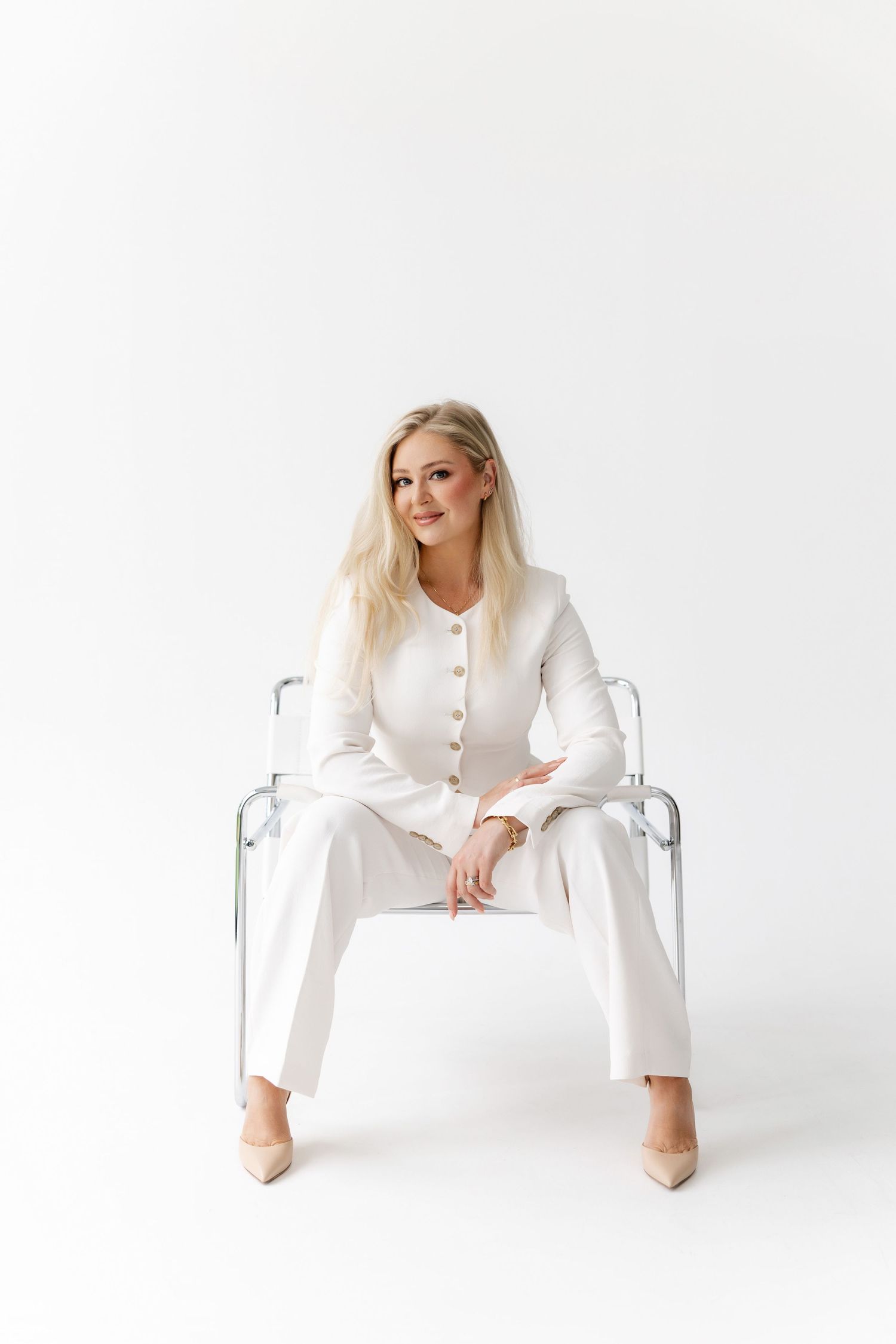 Model in all-white business attire poses confidently on a modern clear chair against white backdrop.
