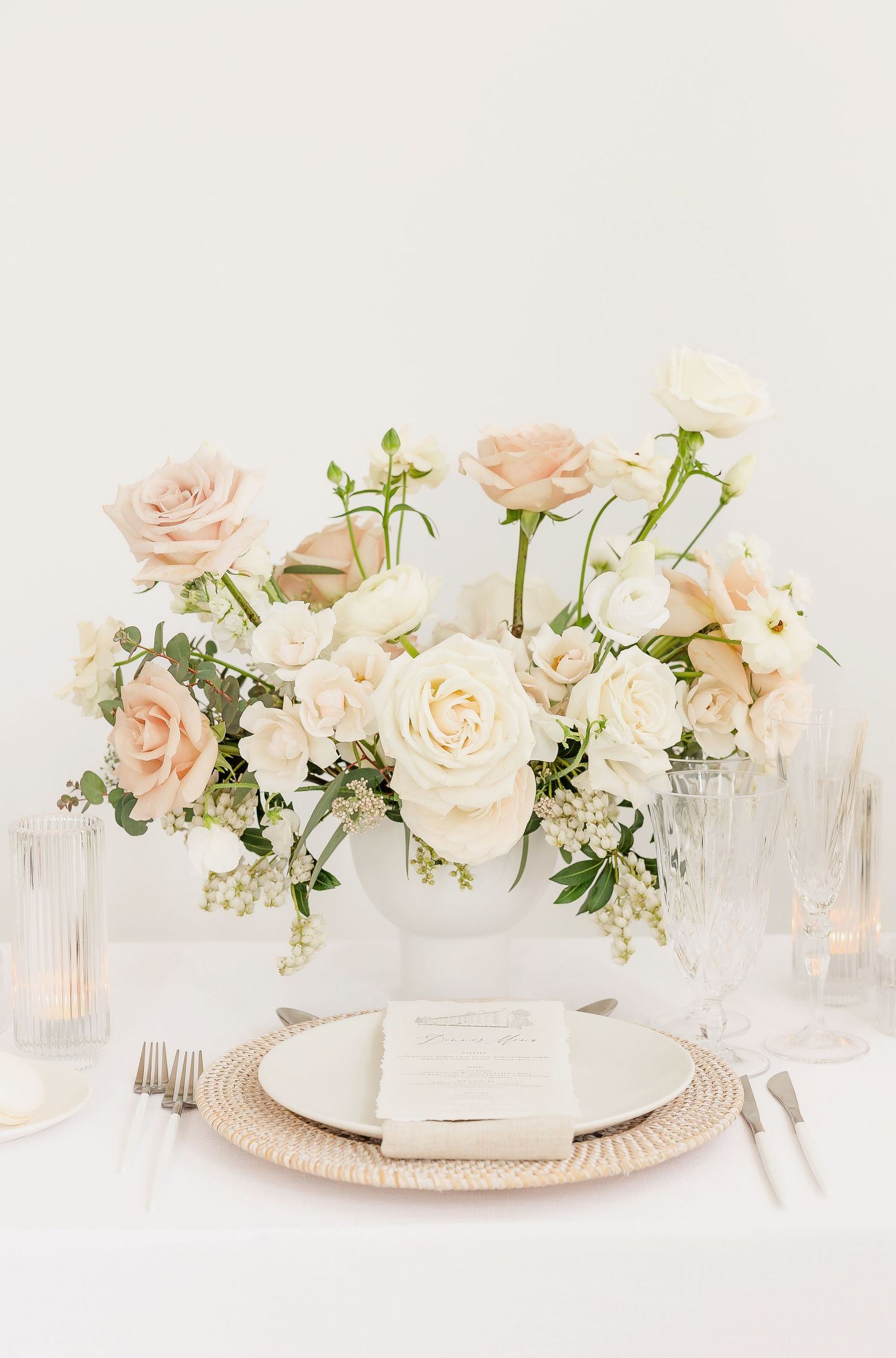 Elegant table setting with blush roses and white floral centerpieces in crystal vases on a light tablecloth.