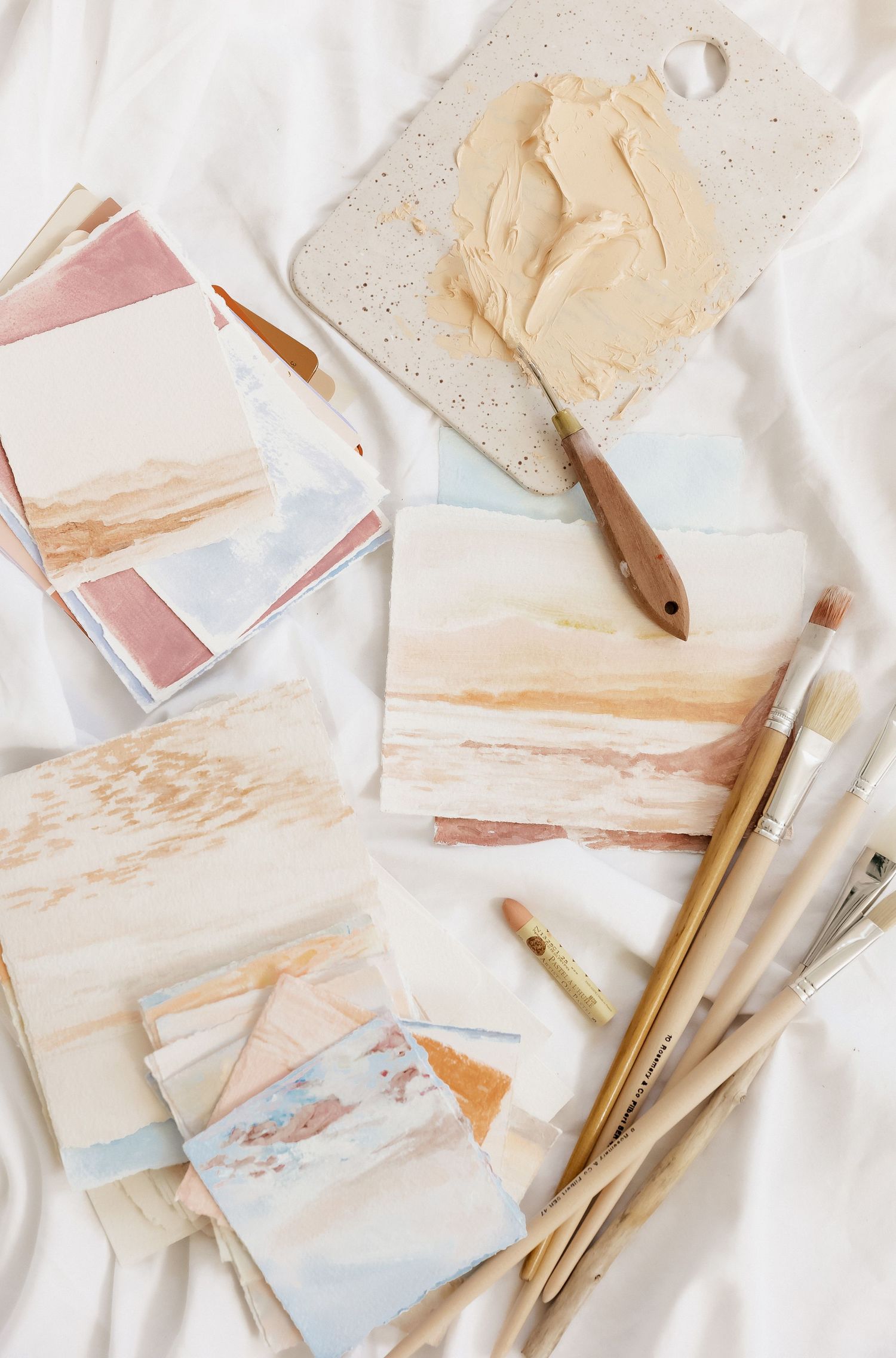 Art supplies and watercolor swatches laid out on white fabric with paintbrushes and palette.