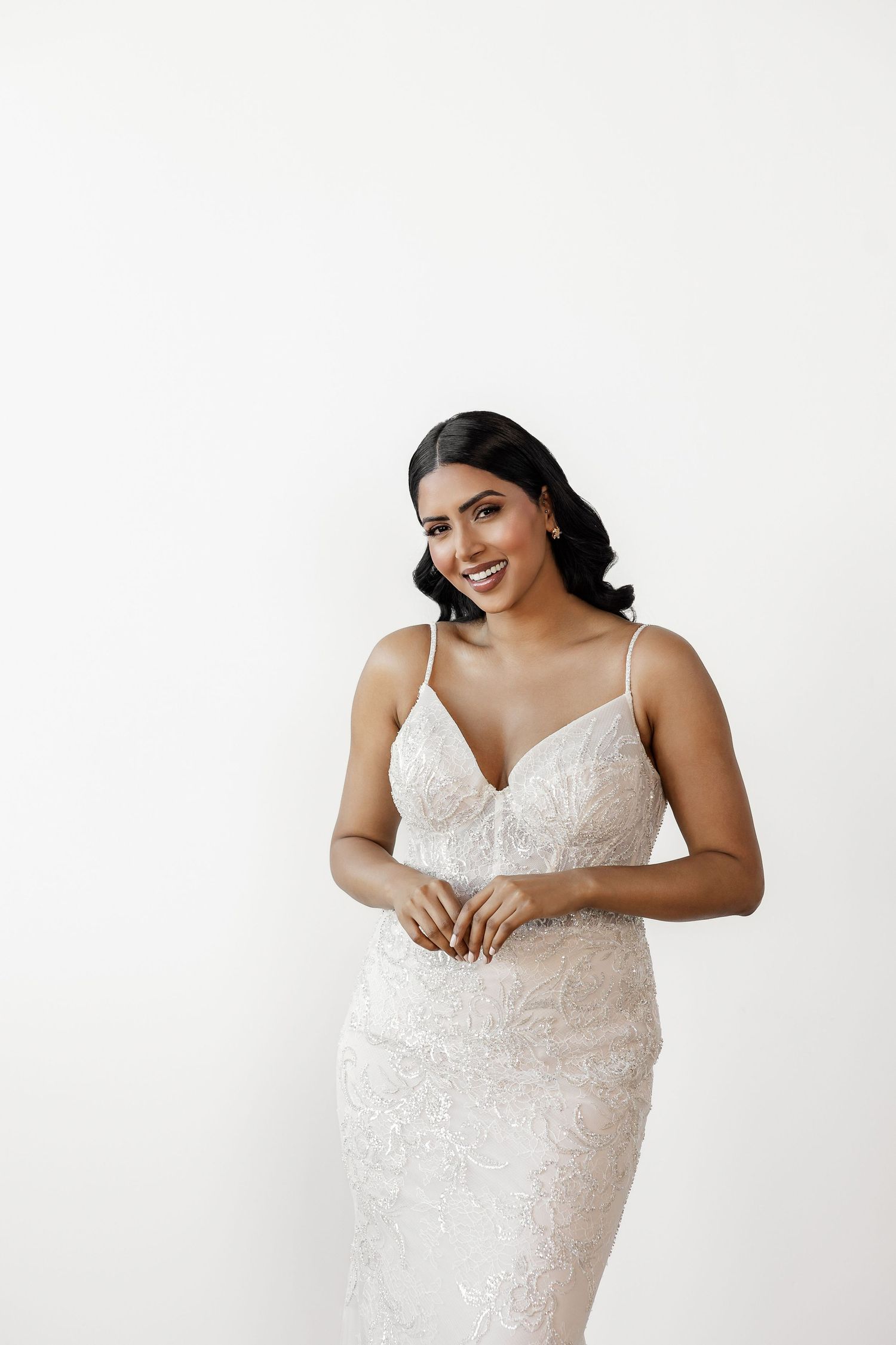 Plus size model poses in a fitted white lace wedding gown against white backdrop.