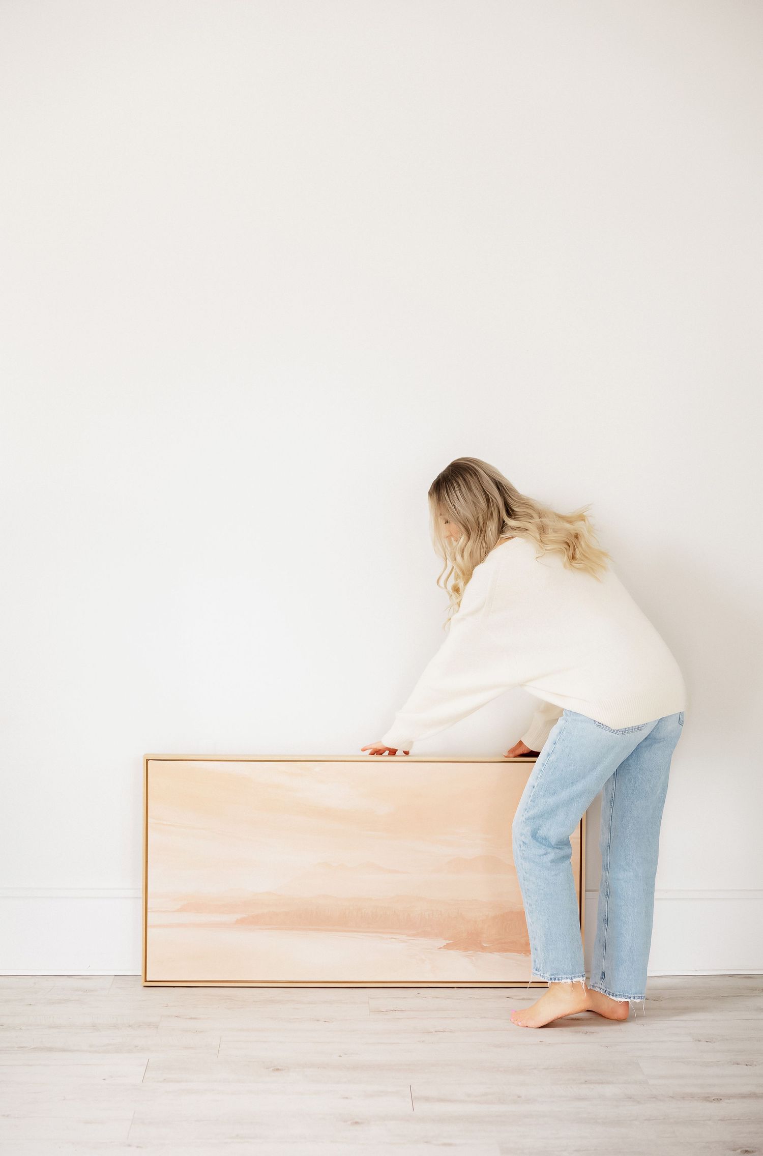 Person in cream sweater and jeans leaning over wooden furniture piece.