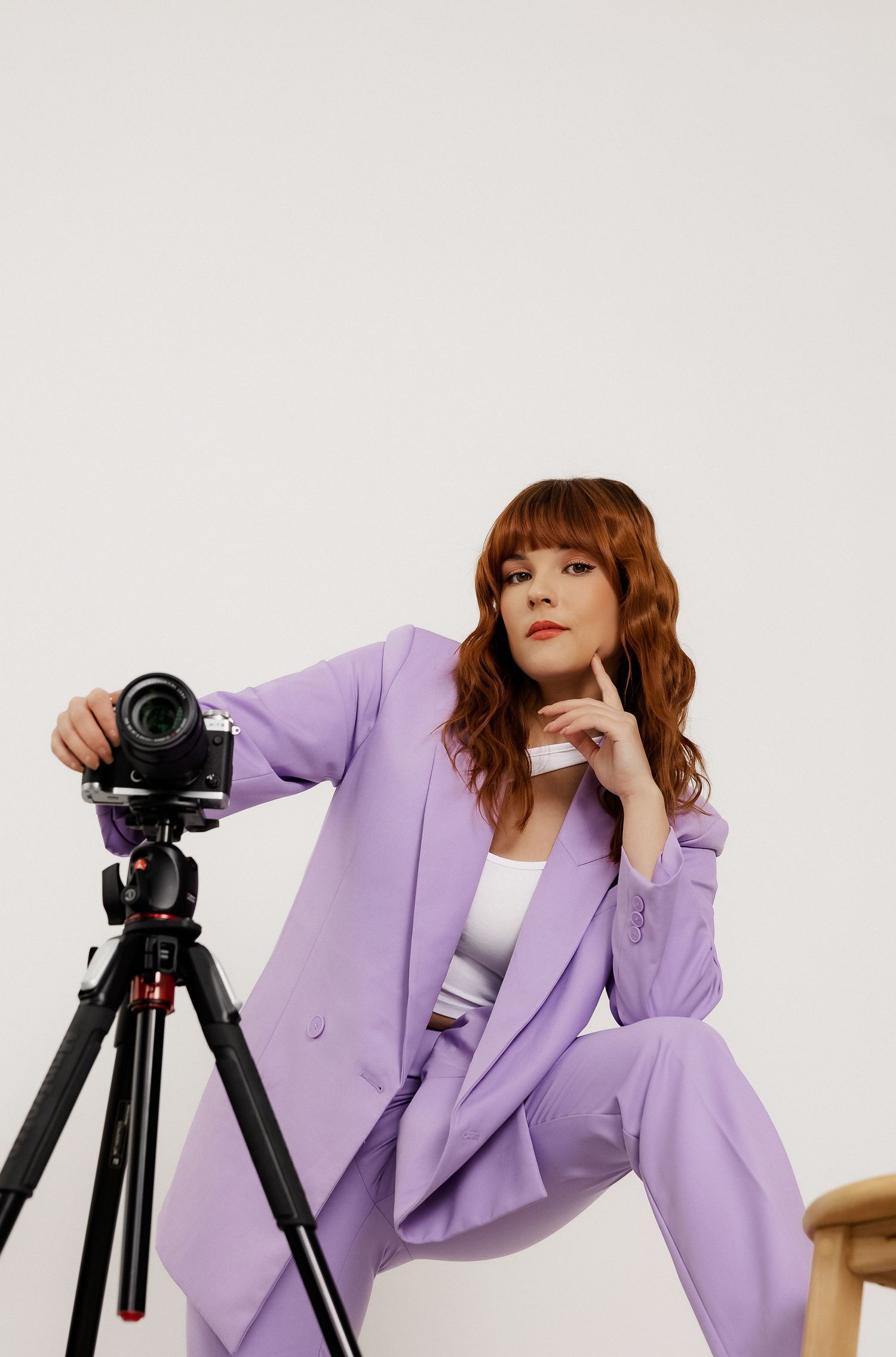 Stylish photographer in purple suit jacket works with camera and tripod in studio setting.