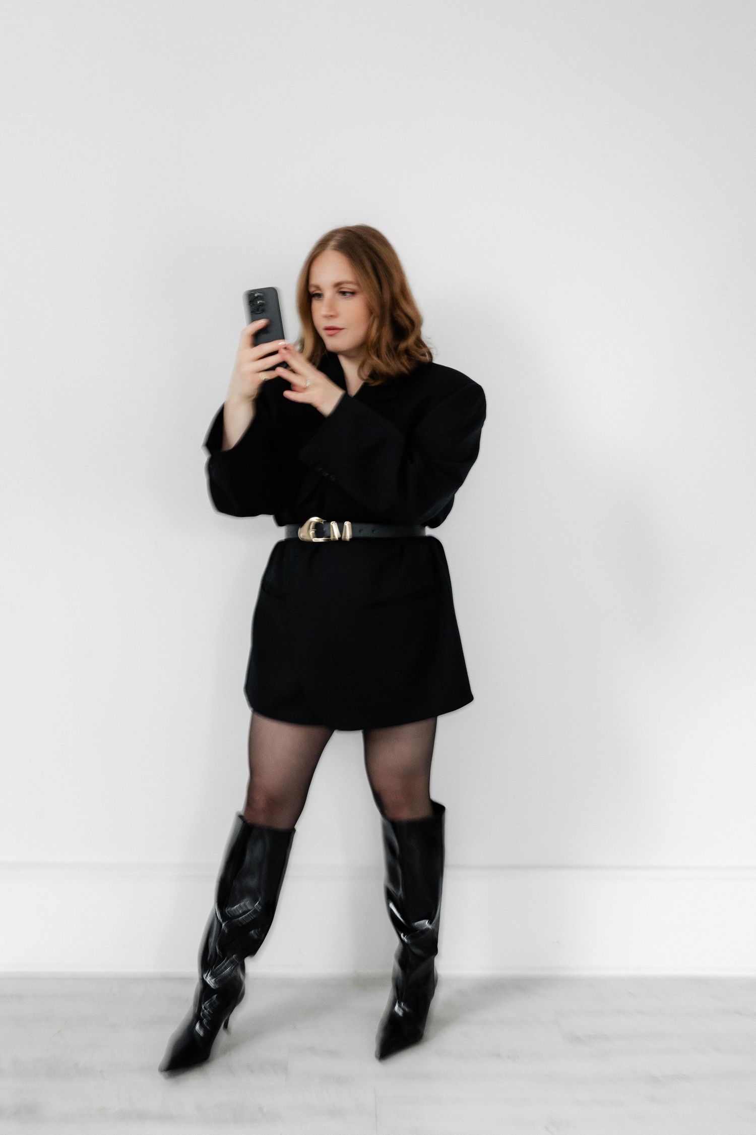 Fashion model in black sweater and boots poses with phone against white backdrop.