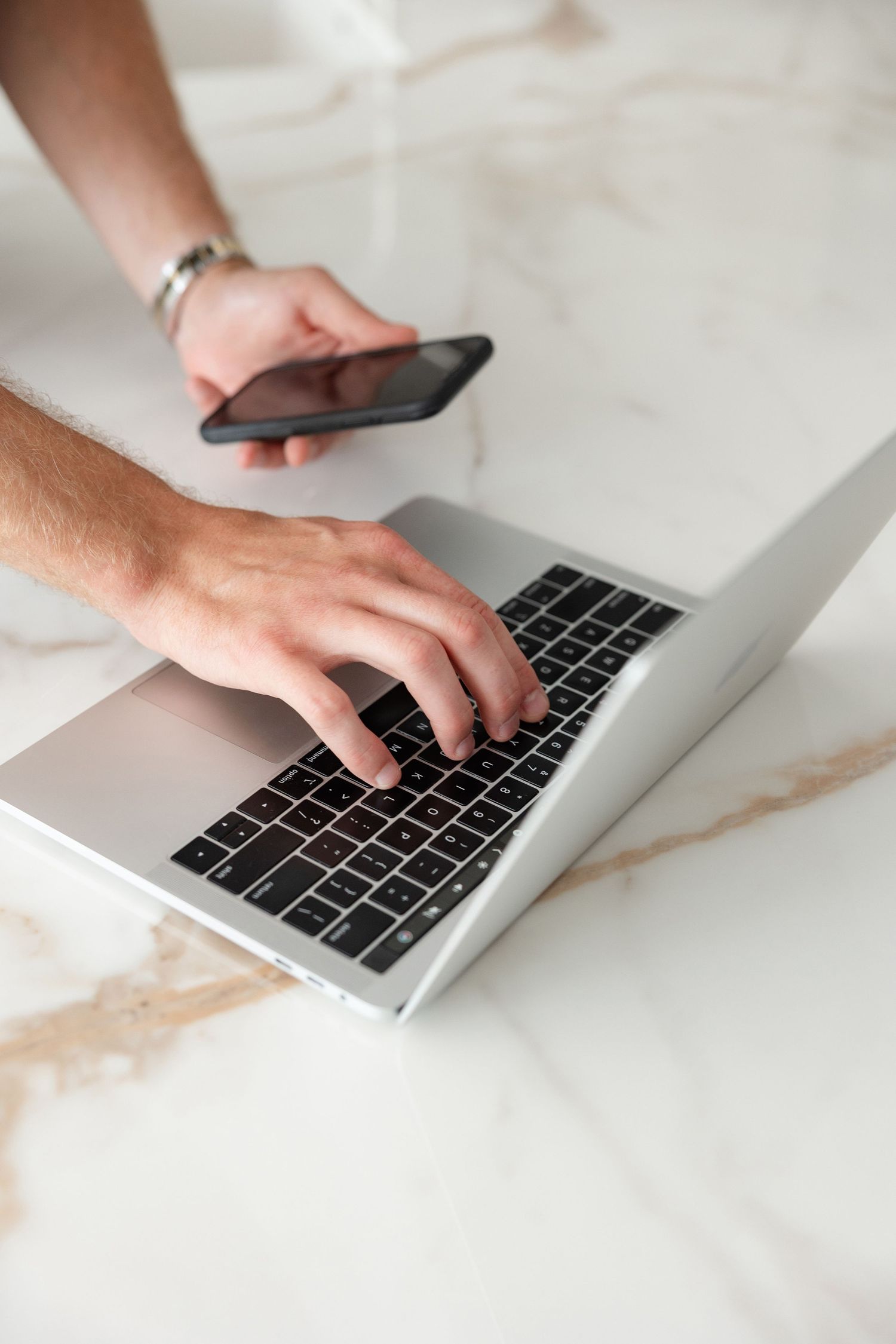 Hands typing on a silver laptop keyboard on a marble countertop.