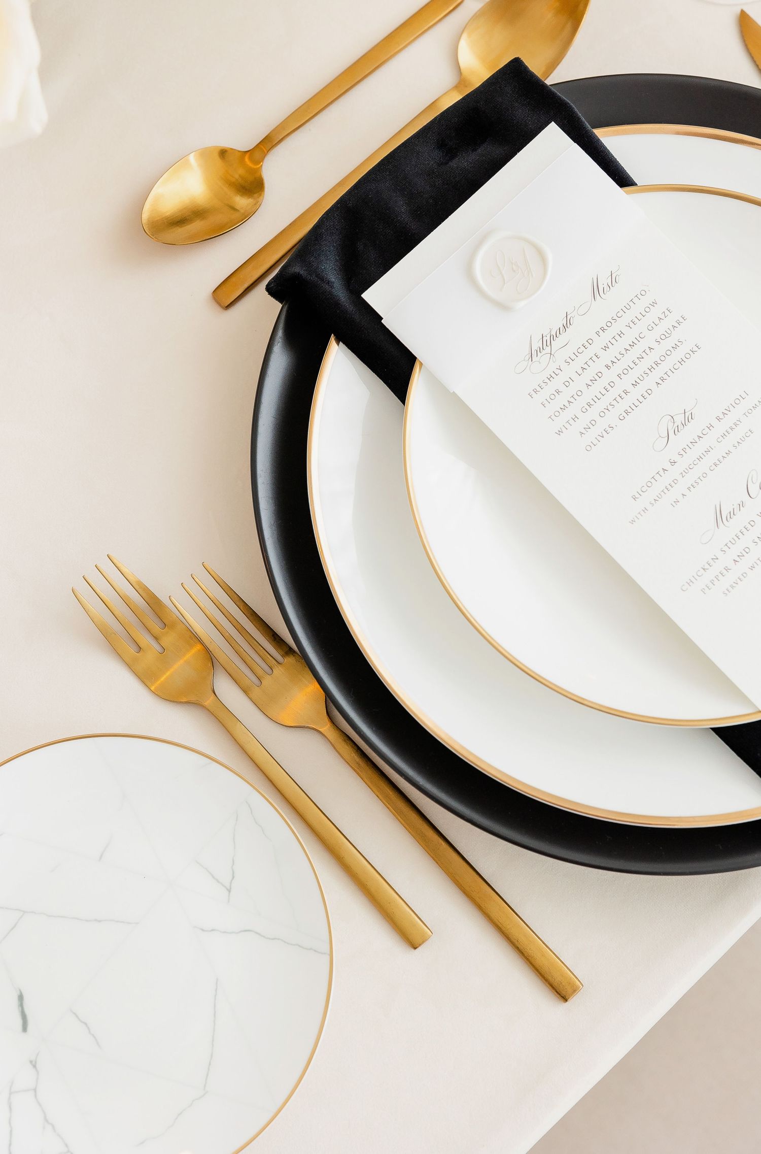 Modern place setting with black charger plate, gold utensils, and white menu card.