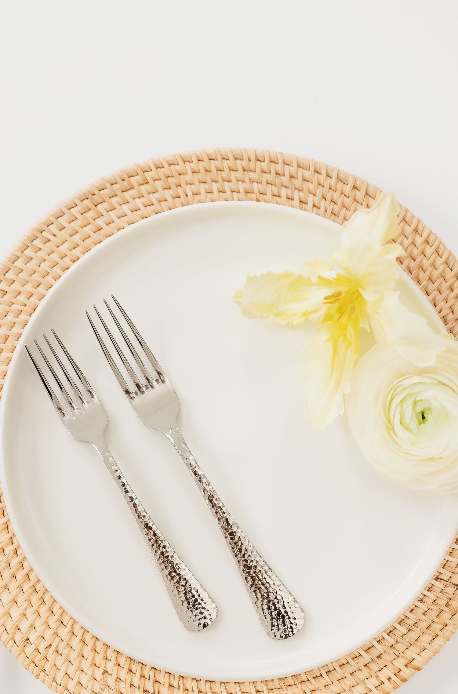 Ornate silver forks on white plate with woven charger and pale yellow flowers.