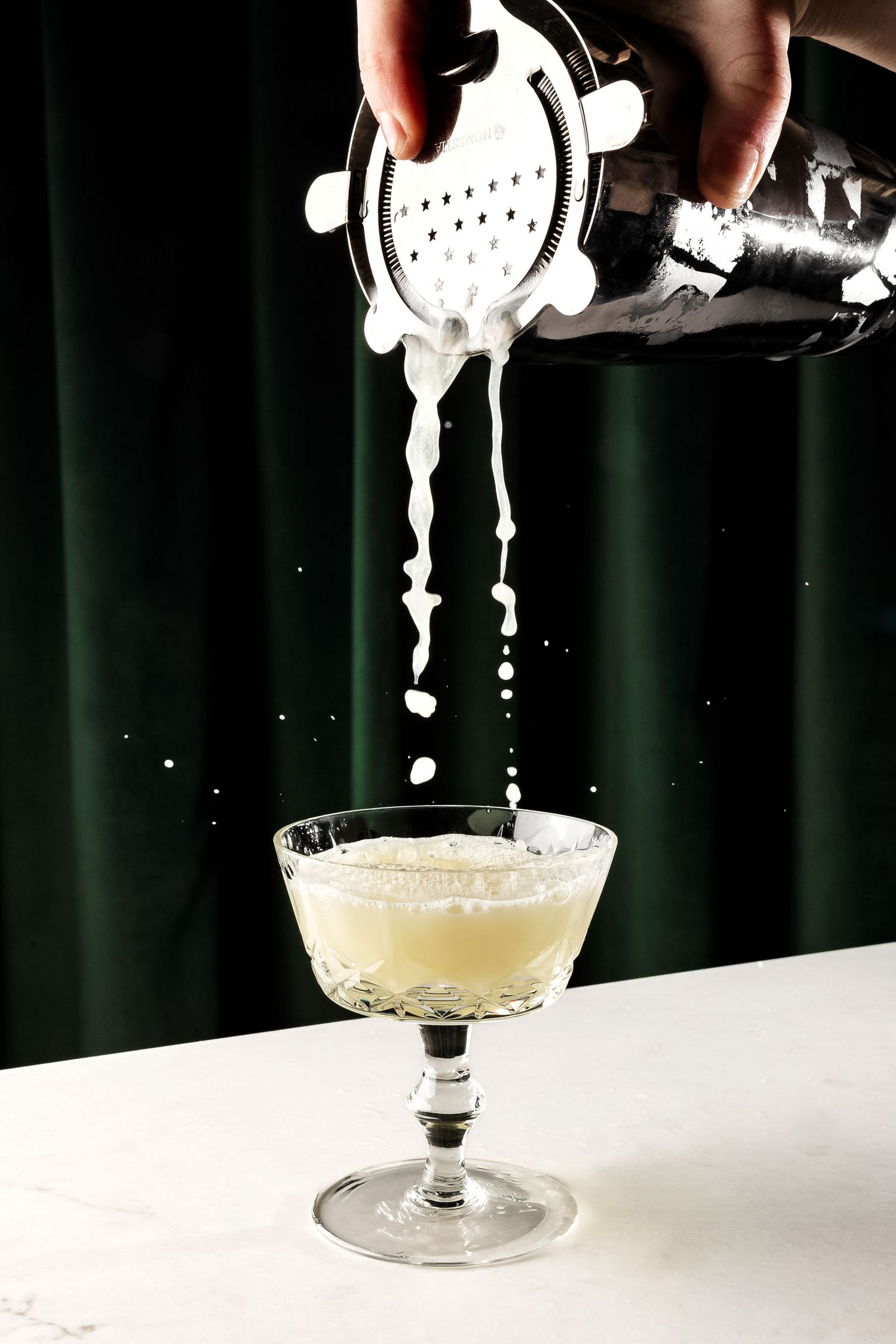White liquid being strained from cocktail shaker into coupe glass.