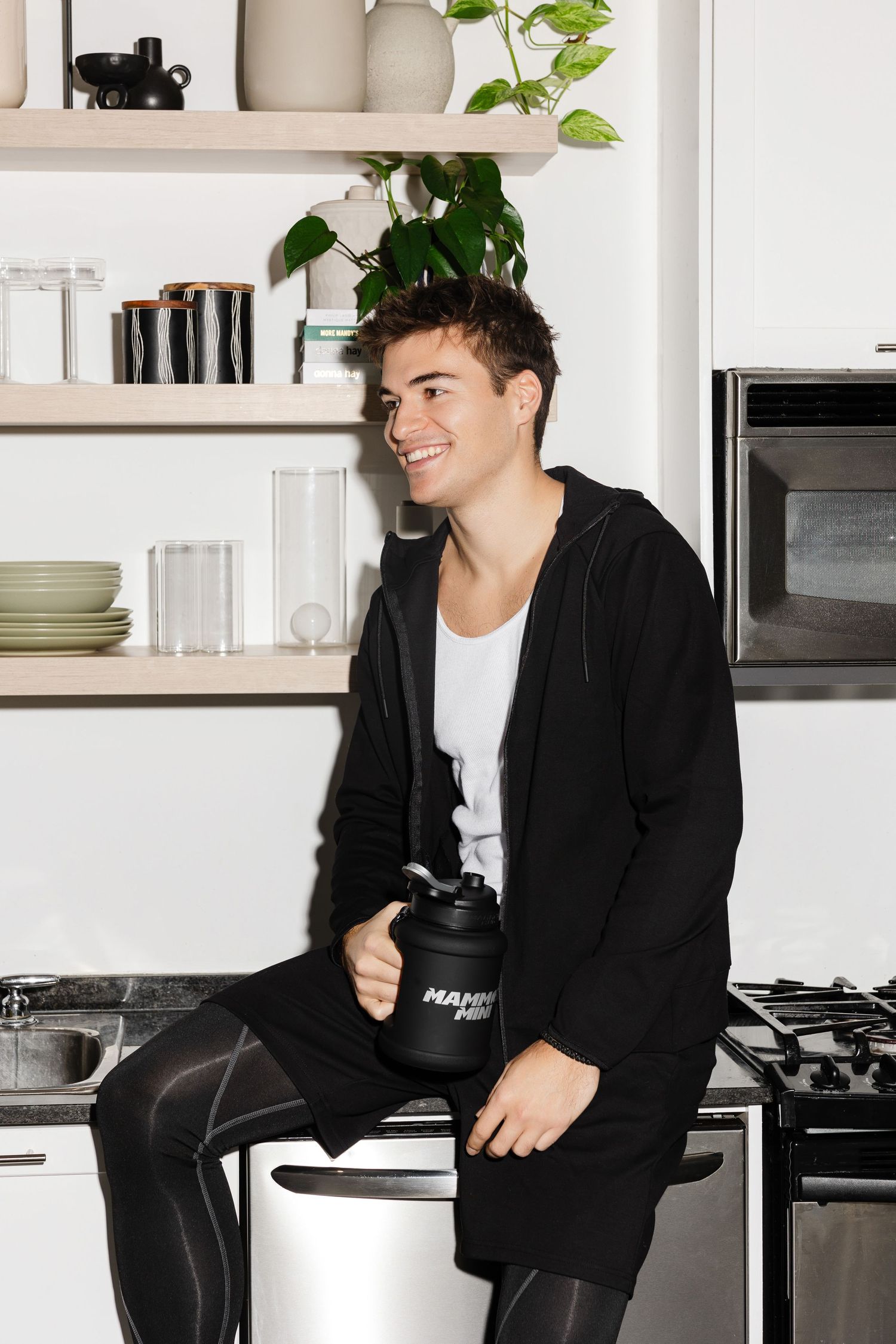 Person in black jacket and white shirt lounges casually in modern white kitchen setting.