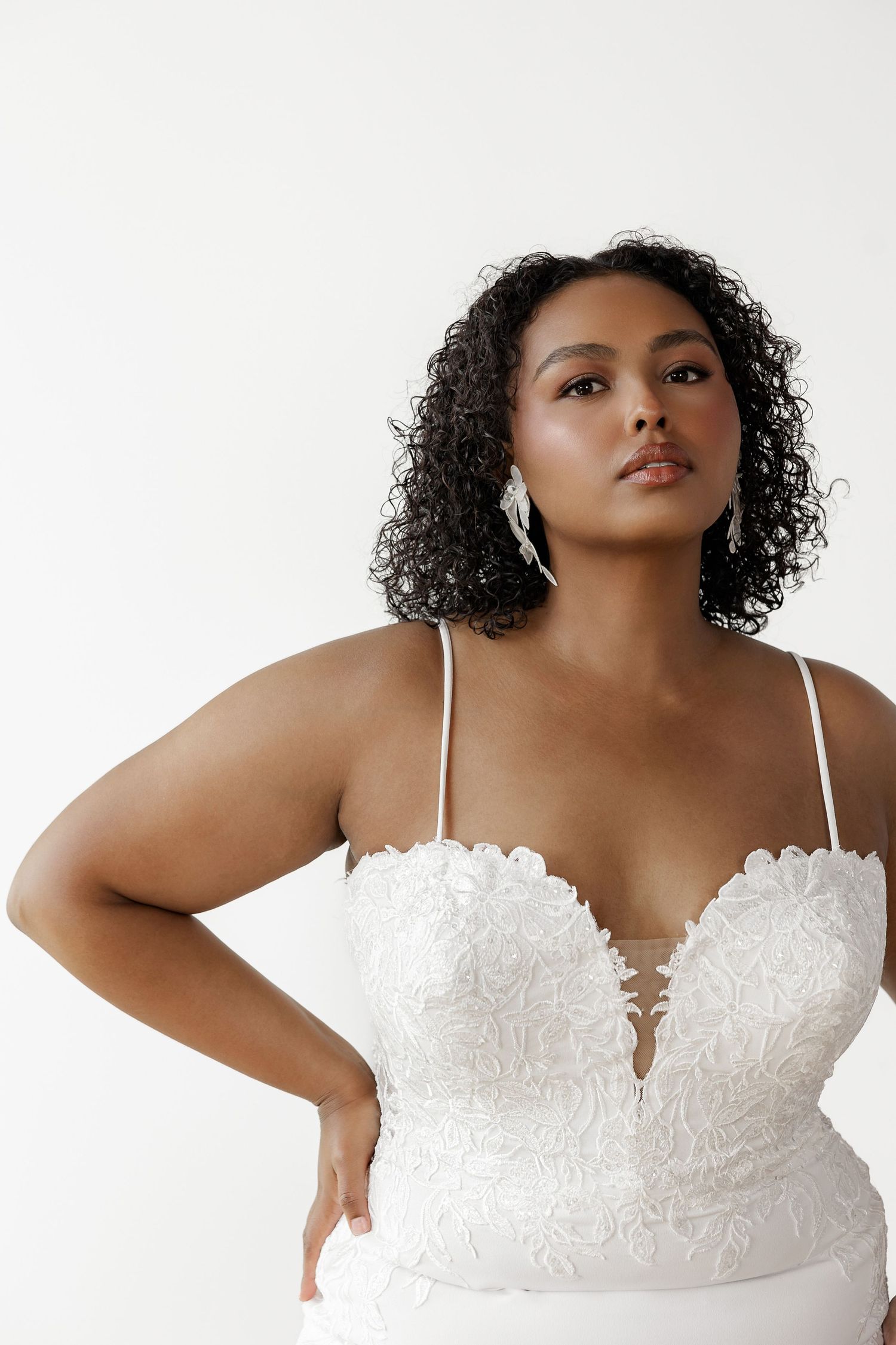 Plus size model showcases elegant white lace bodice wedding dress against studio backdrop.