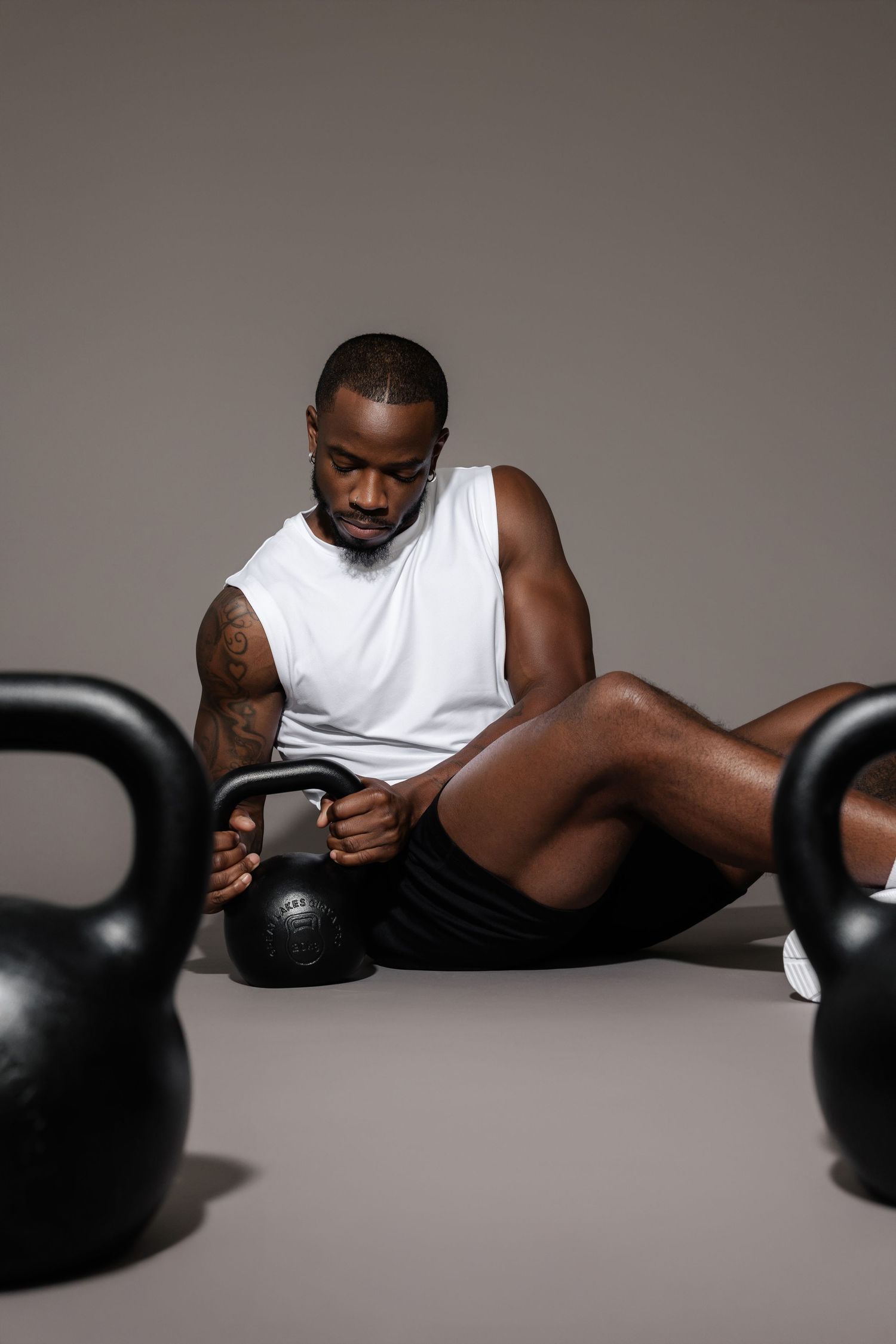 Athletic figure in white tank top lying on floor between black kettlebells.
