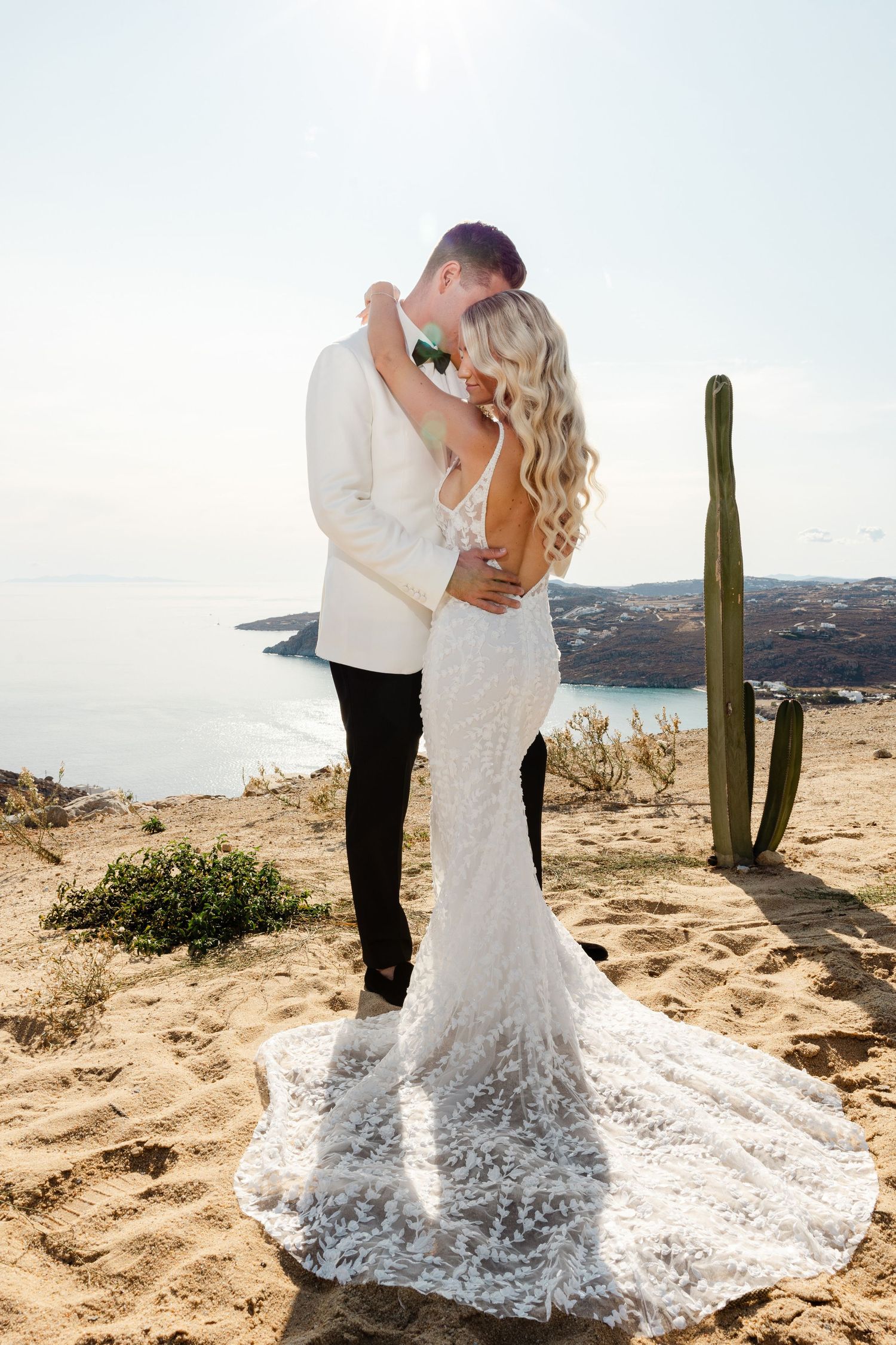 A romantic wedding couple embraces on a desert cliff with cacti and ocean views in the background.