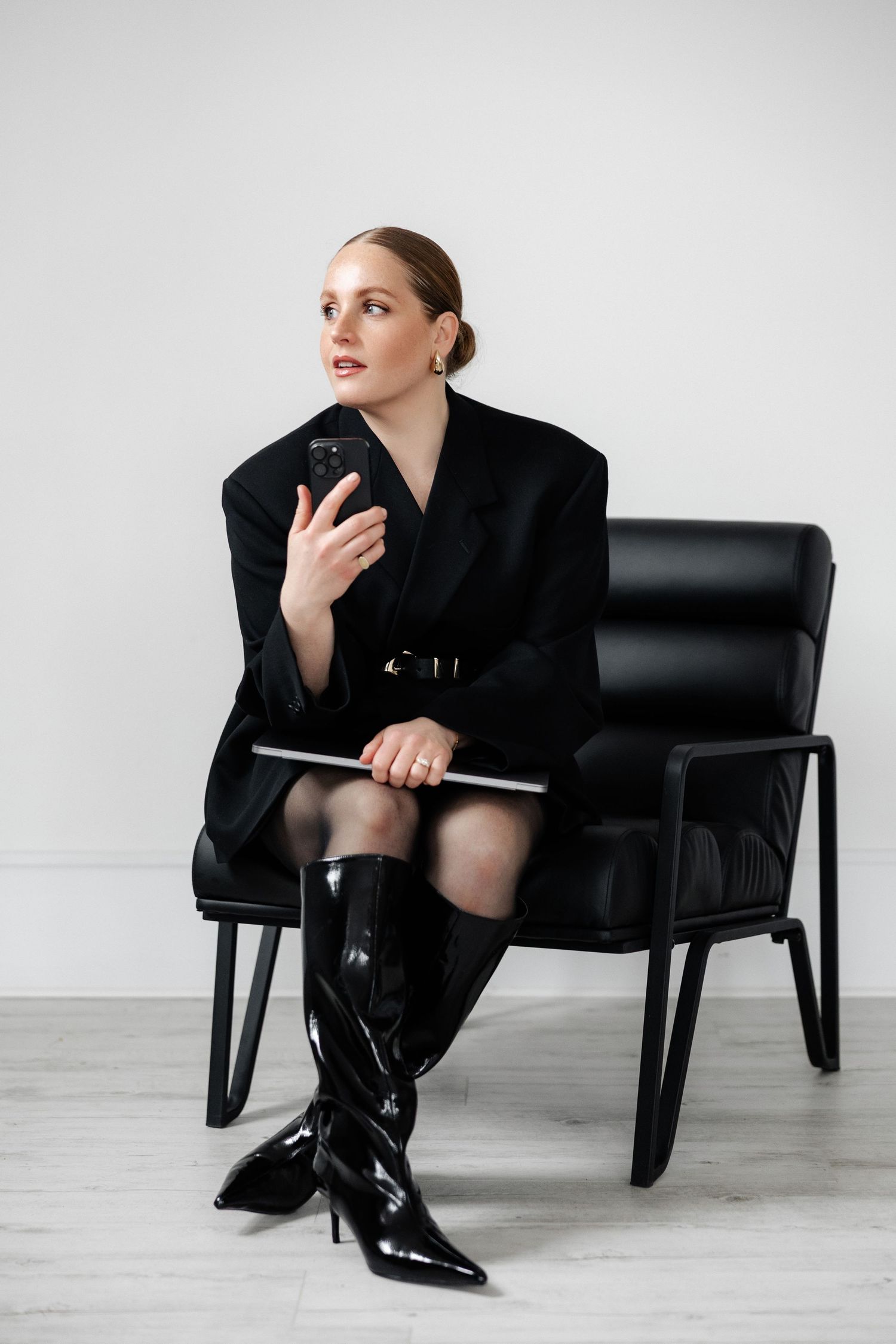 Professional wearing black blazer and knee-high boots sits in leather chair while holding smartphone.