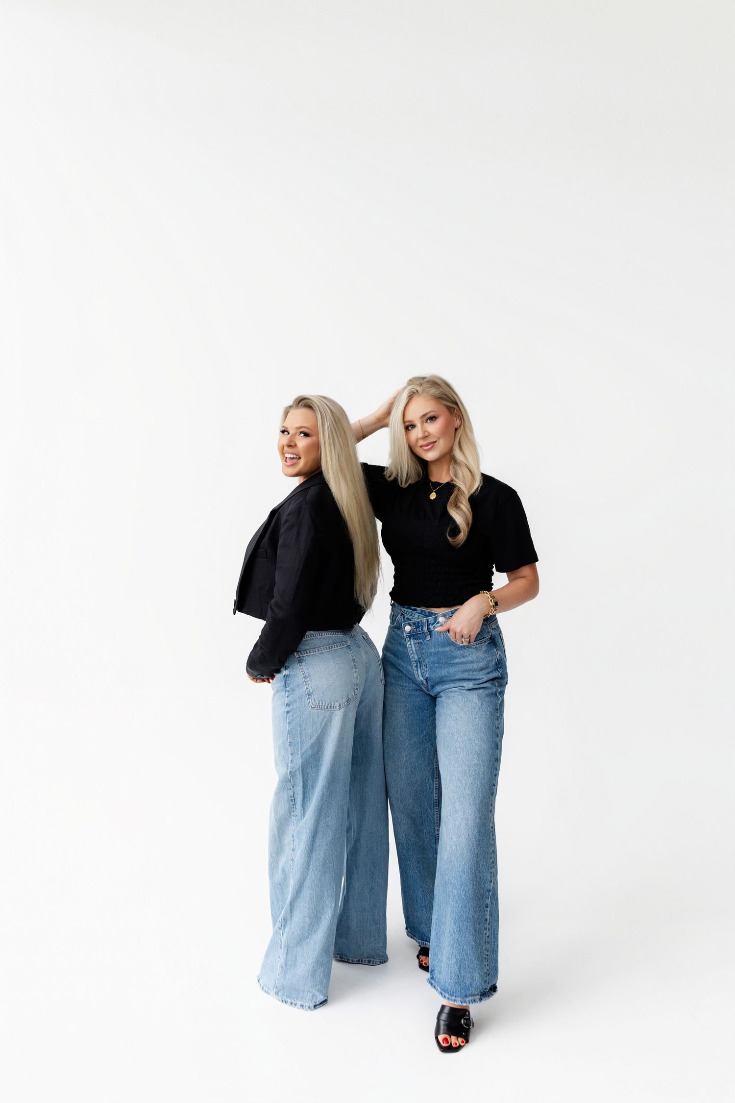 Two people in black tops and light denim jeans posing against white background.