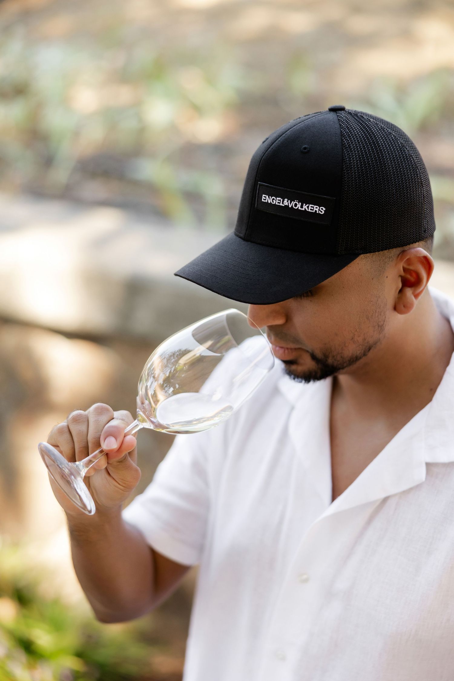 Person in white shirt and black cap tasting wine outdoors.